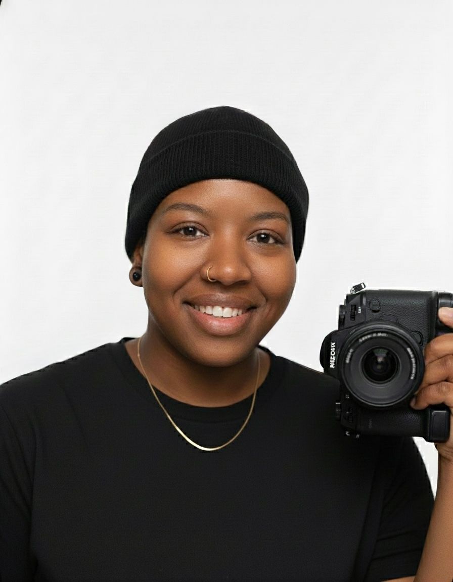 Smiling photographer wearing a black beanie and holding a DSLR camera against a simple white background.