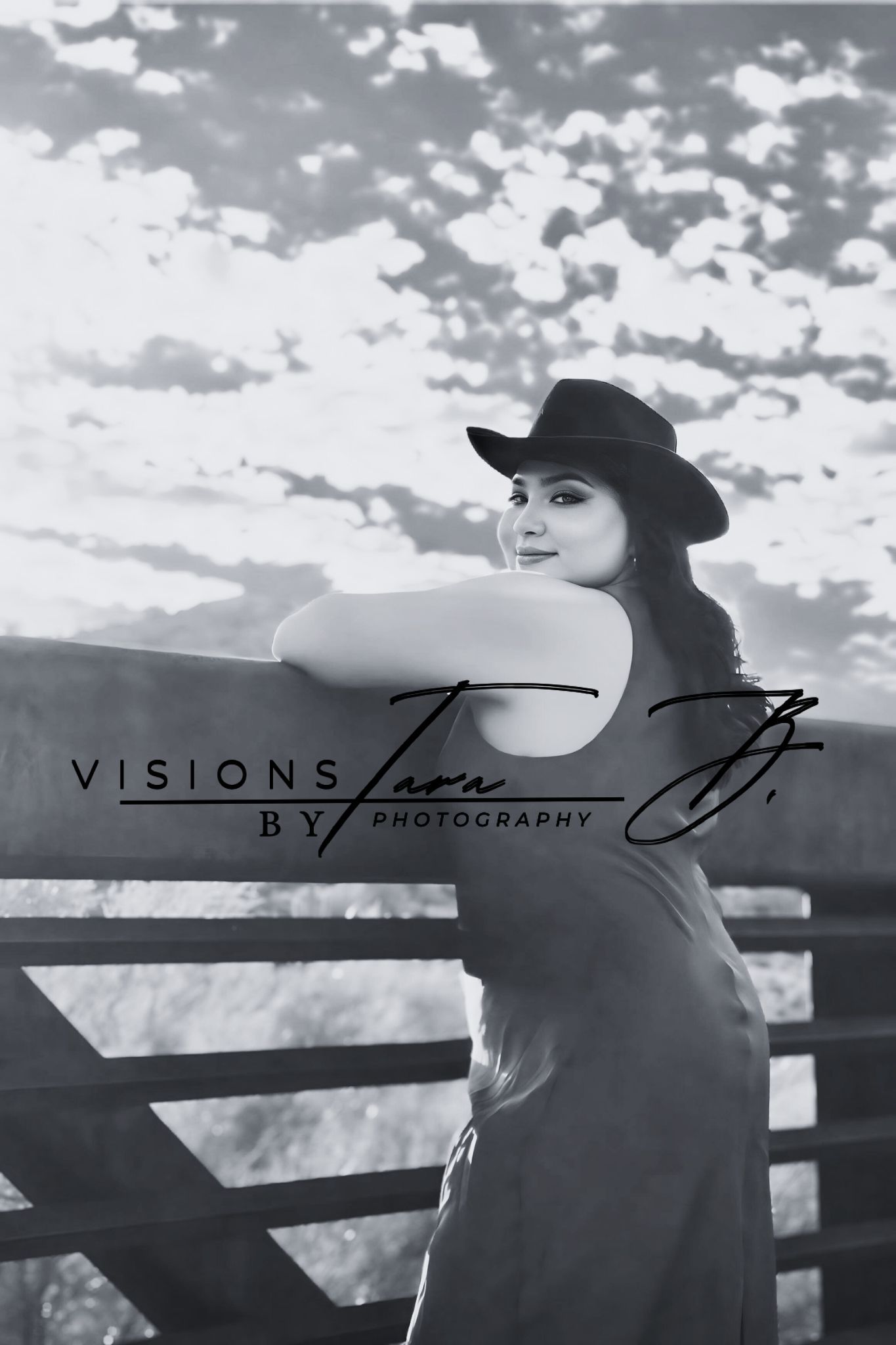 Black-and-white portrait of a woman in a cowboy hat leaning on a wooden fence, captured against a dramatic sky.