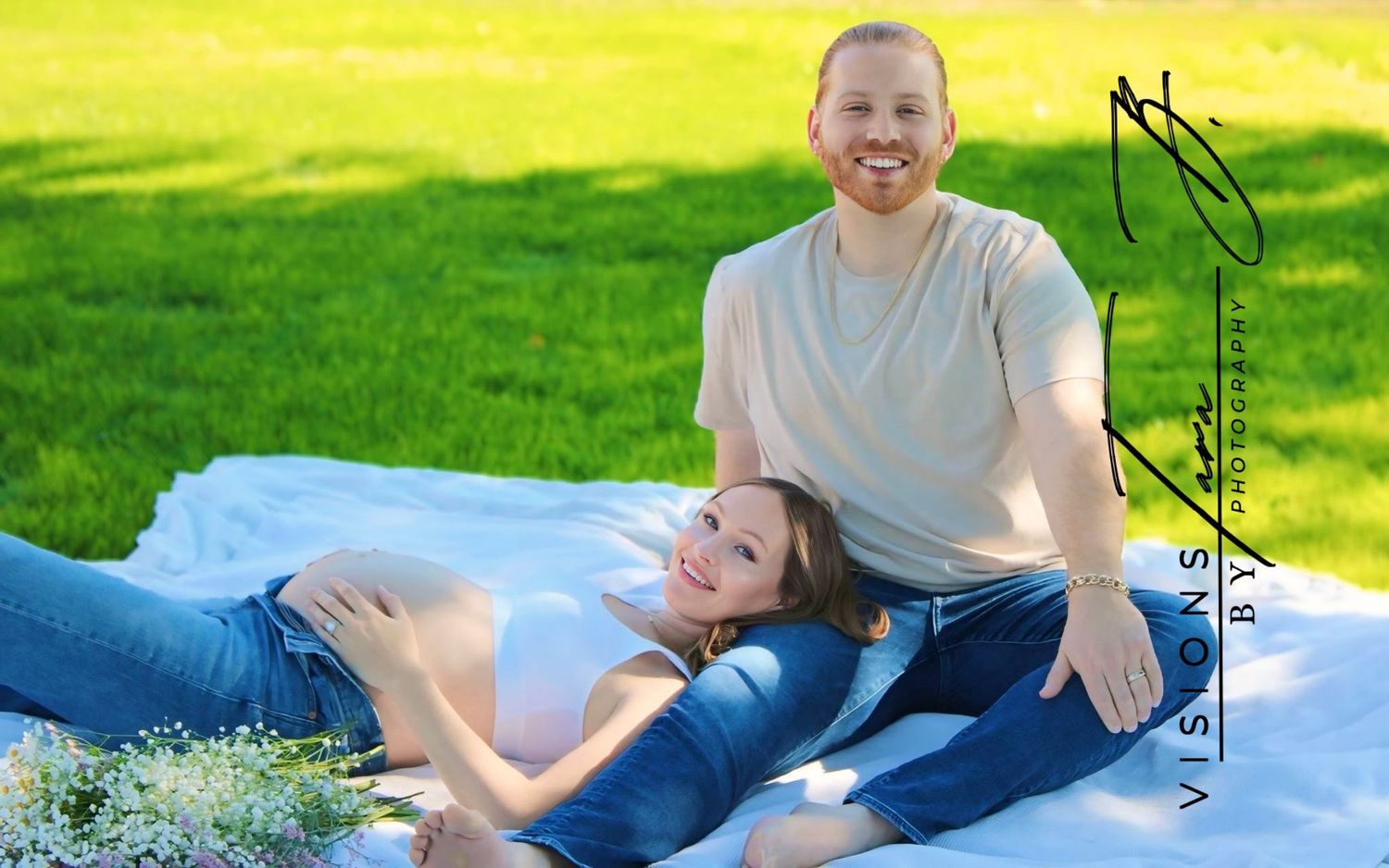 Pregnant couple laughing together on a picnic blanket in a sunny field, enjoying a relaxed and joyful moment outdoors.