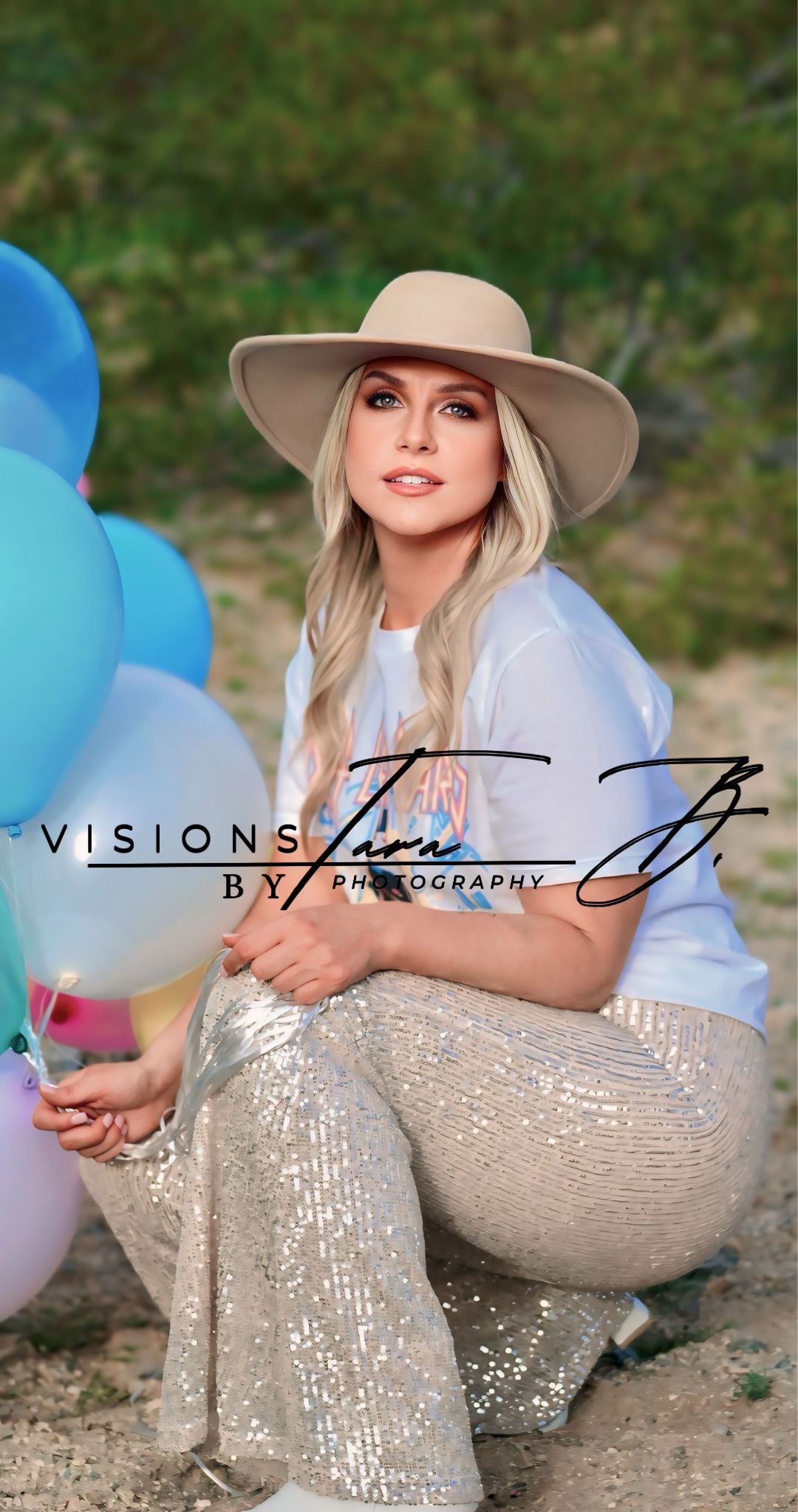 Woman sitting in the desert holding colorful balloons, smiling softly while dressed in a stylish hat and neutral outfit.