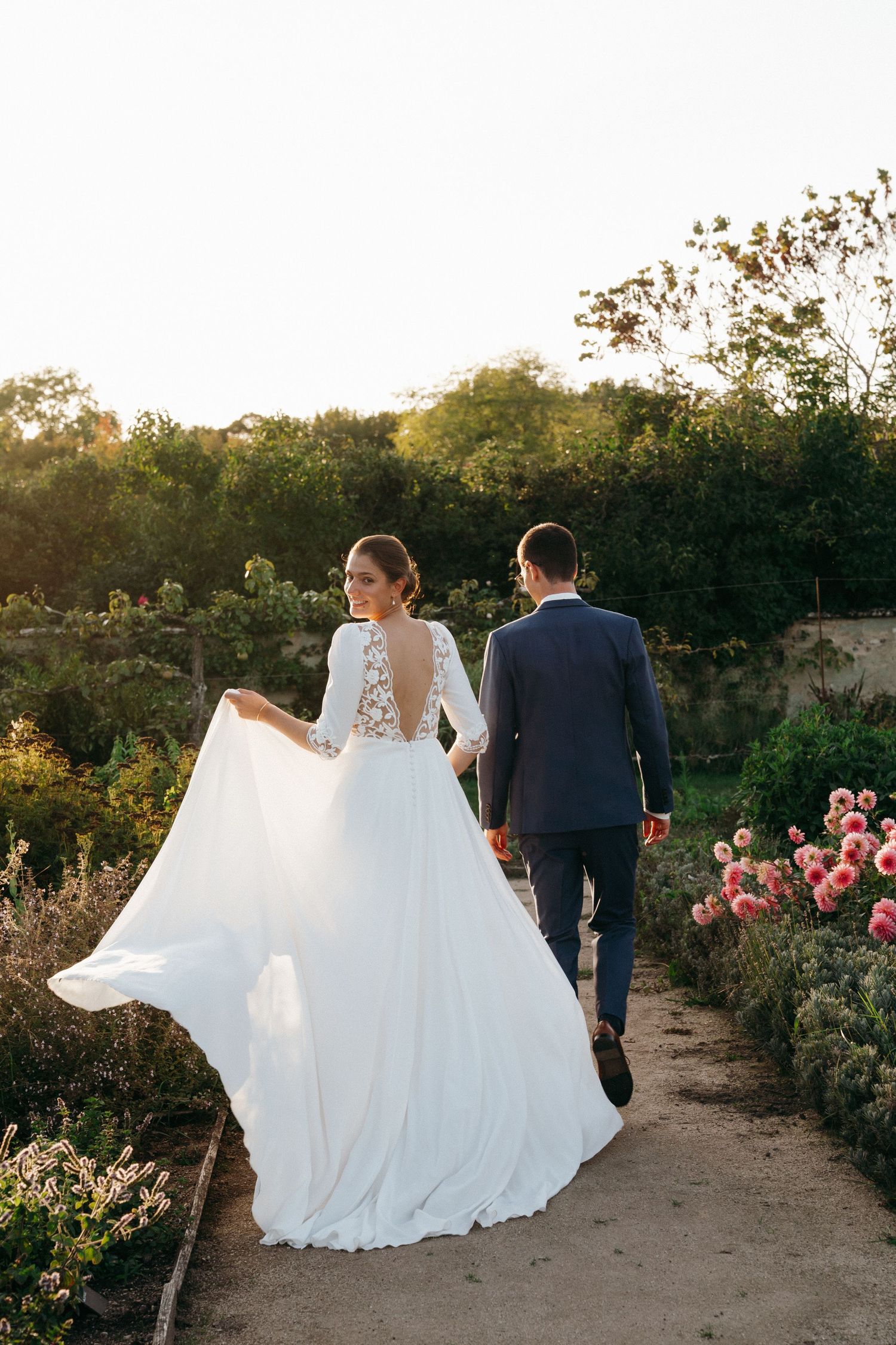 Bride in elegant backless lace wedding gown walks with groom through a blooming garden at sunset.