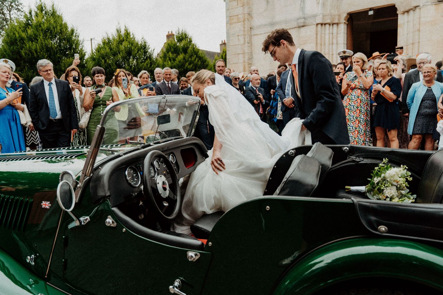 Mariés sortant d'une voiture ancienne verte, entourés d'invités après leur messe de mariage