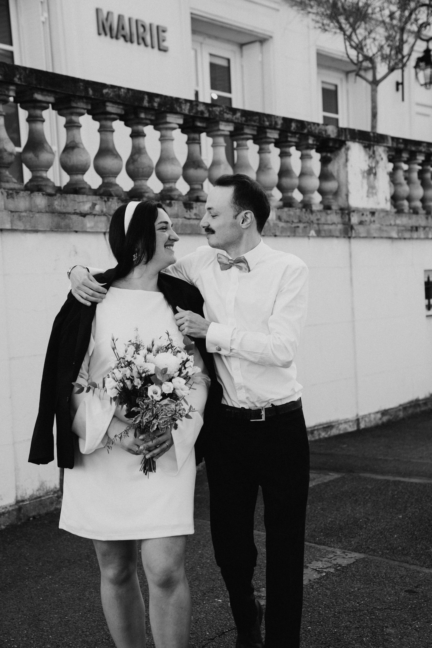Black and white photo of newlywed couple embracing outside a French Mairie with bouquet.
