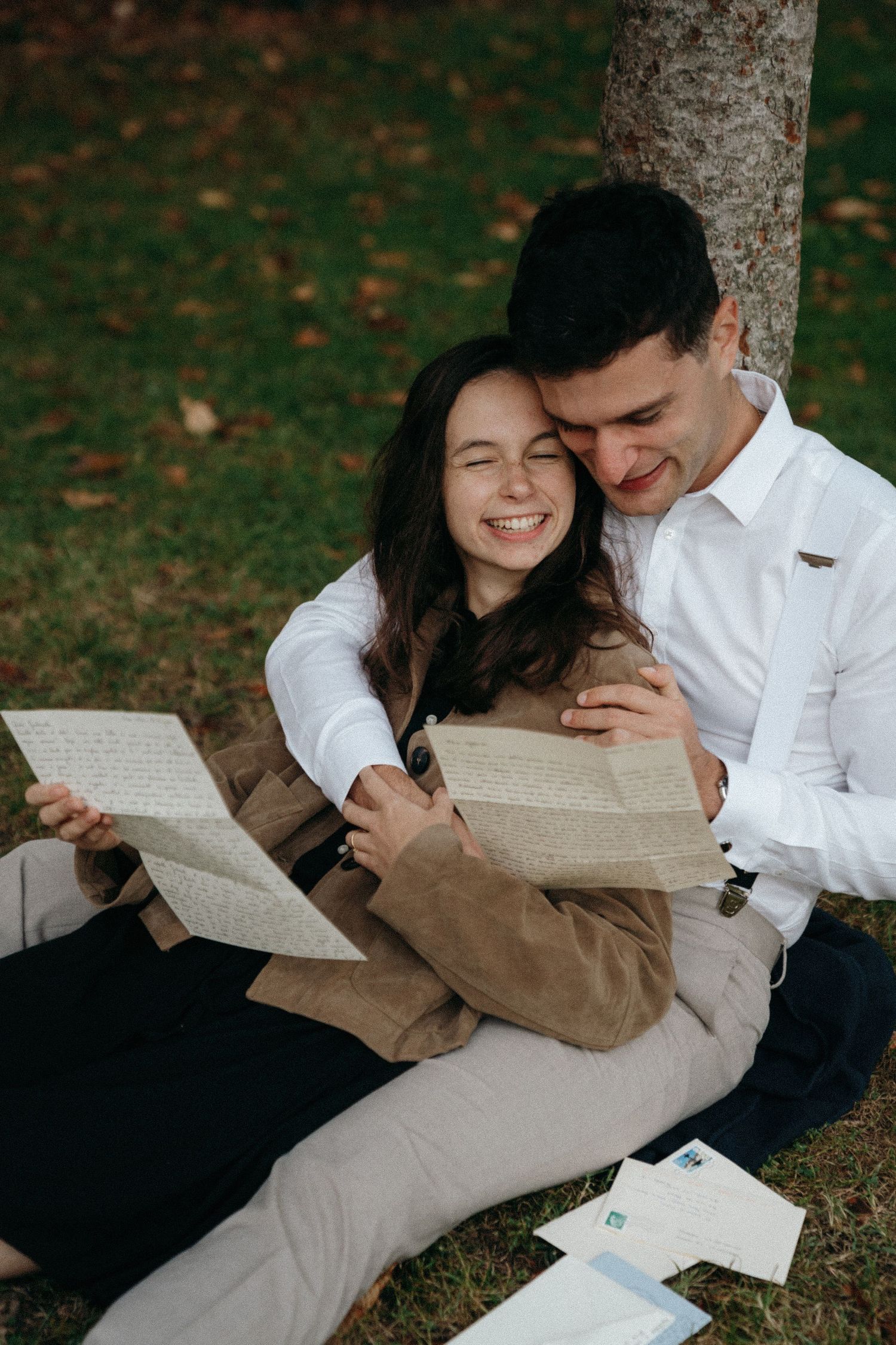 Happy couple sitting against tree in park, smiling while reading handwritten love letters together.