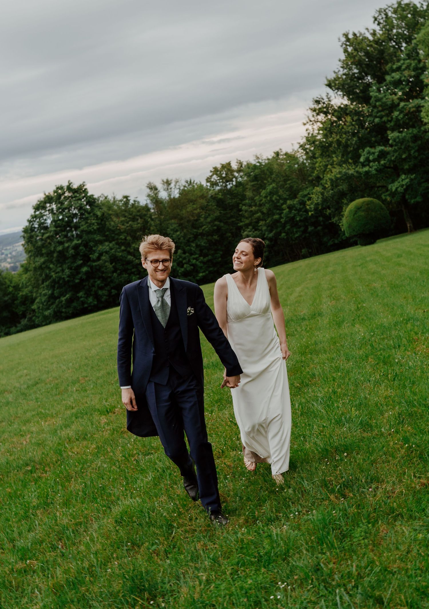 Bride and groom walk hand in hand across a green hillside meadow under a cloudy sky.