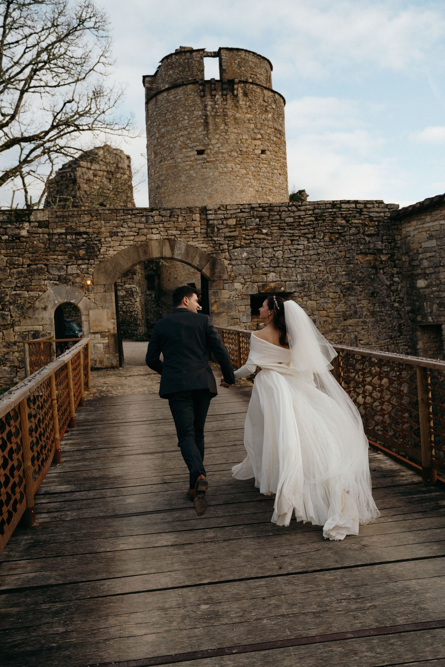 Le jour de leur mariage, les mariés, main dans la main, traversent un pont en bois en direction de la tour d'un château en pierre.