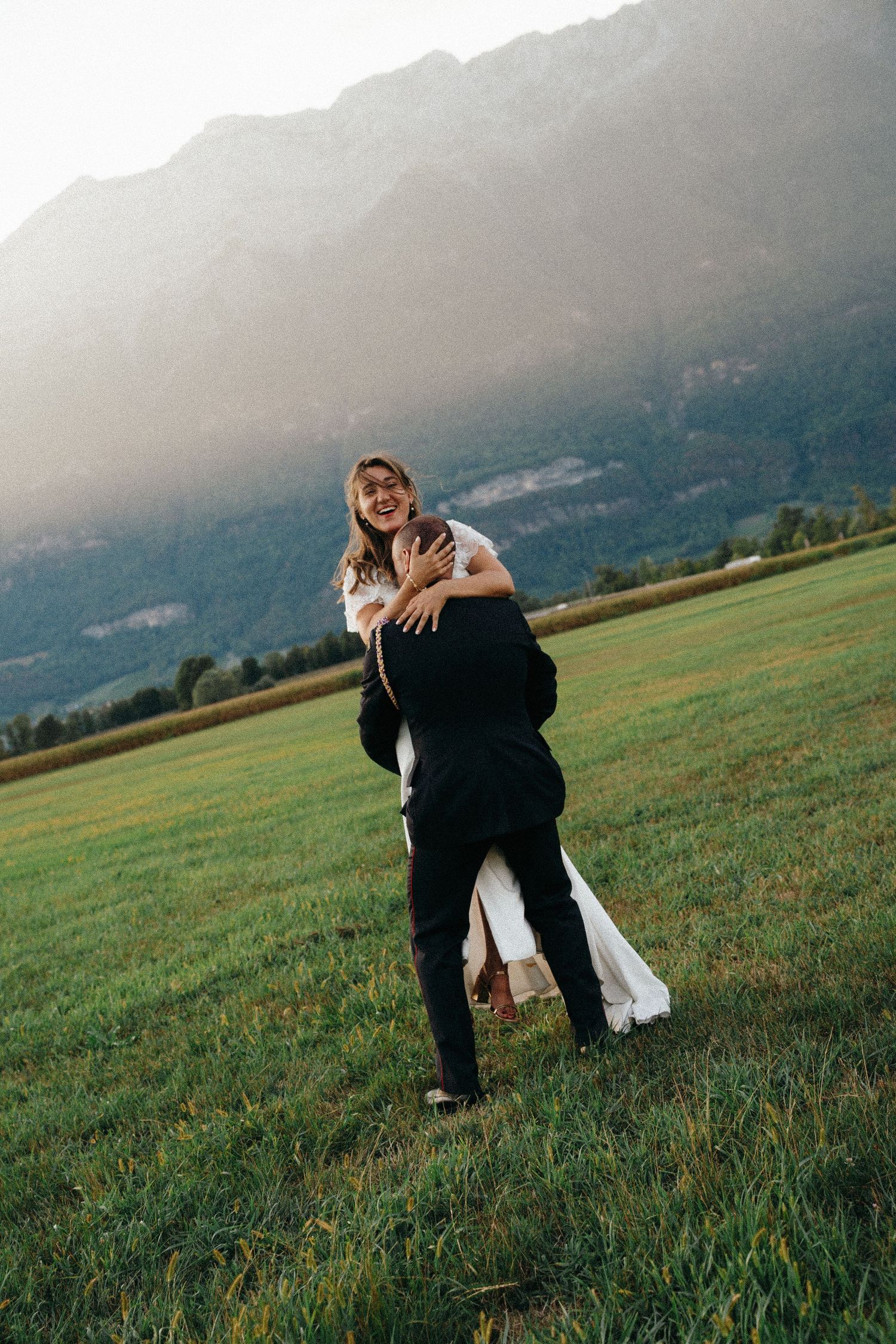 Joyful groom lifts laughing bride in white dress in a green meadow with mountain backdrop.