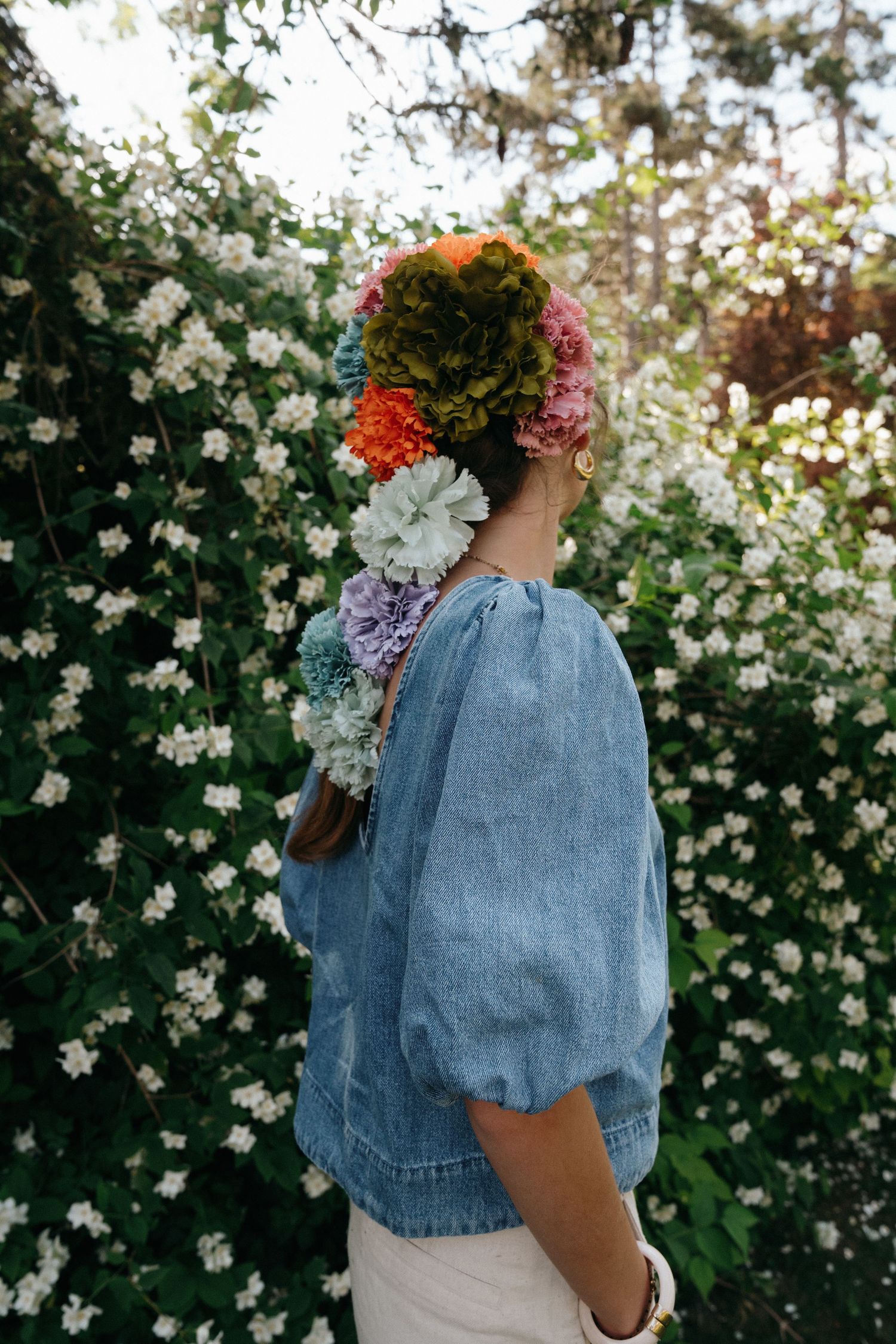 Woman in denim blouse with colorful flower crown standing before blooming white bushes.
