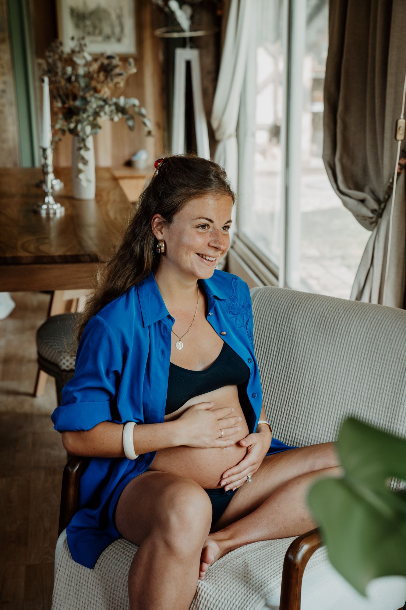 Smiling pregnant woman in blue shirt cradling baby bump seated in cozy armchair.