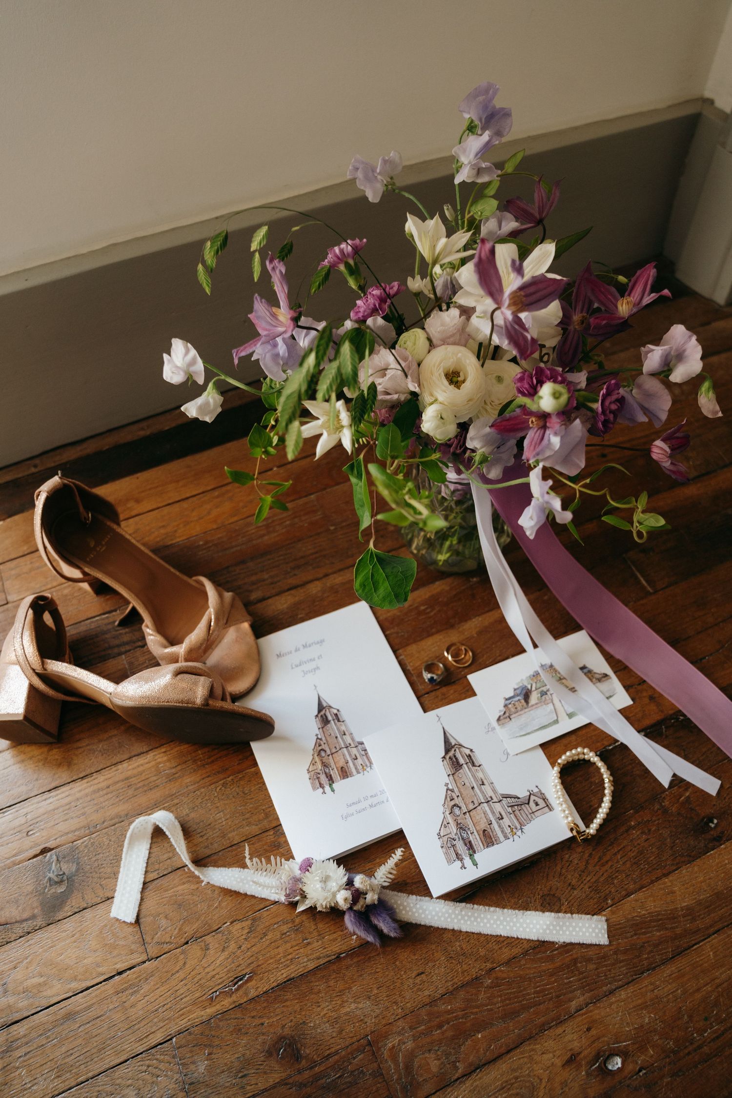 Elegant wedding flat lay featuring purple floral bouquet, invitation cards, gold rings, and blush heels on wood floor.