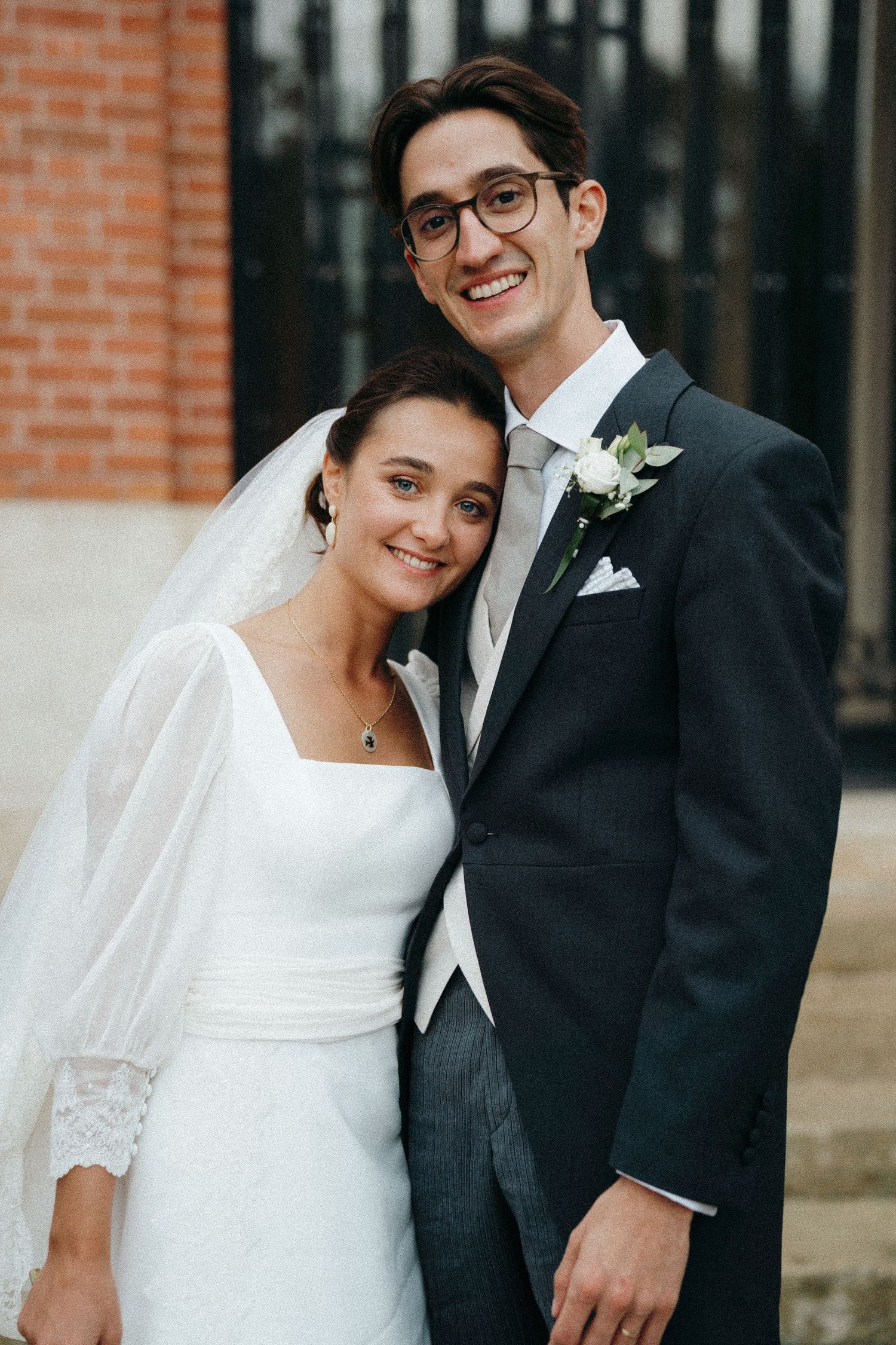 Smiling bride in white gown with veil and groom in suit with boutonniere pose together outdoors.