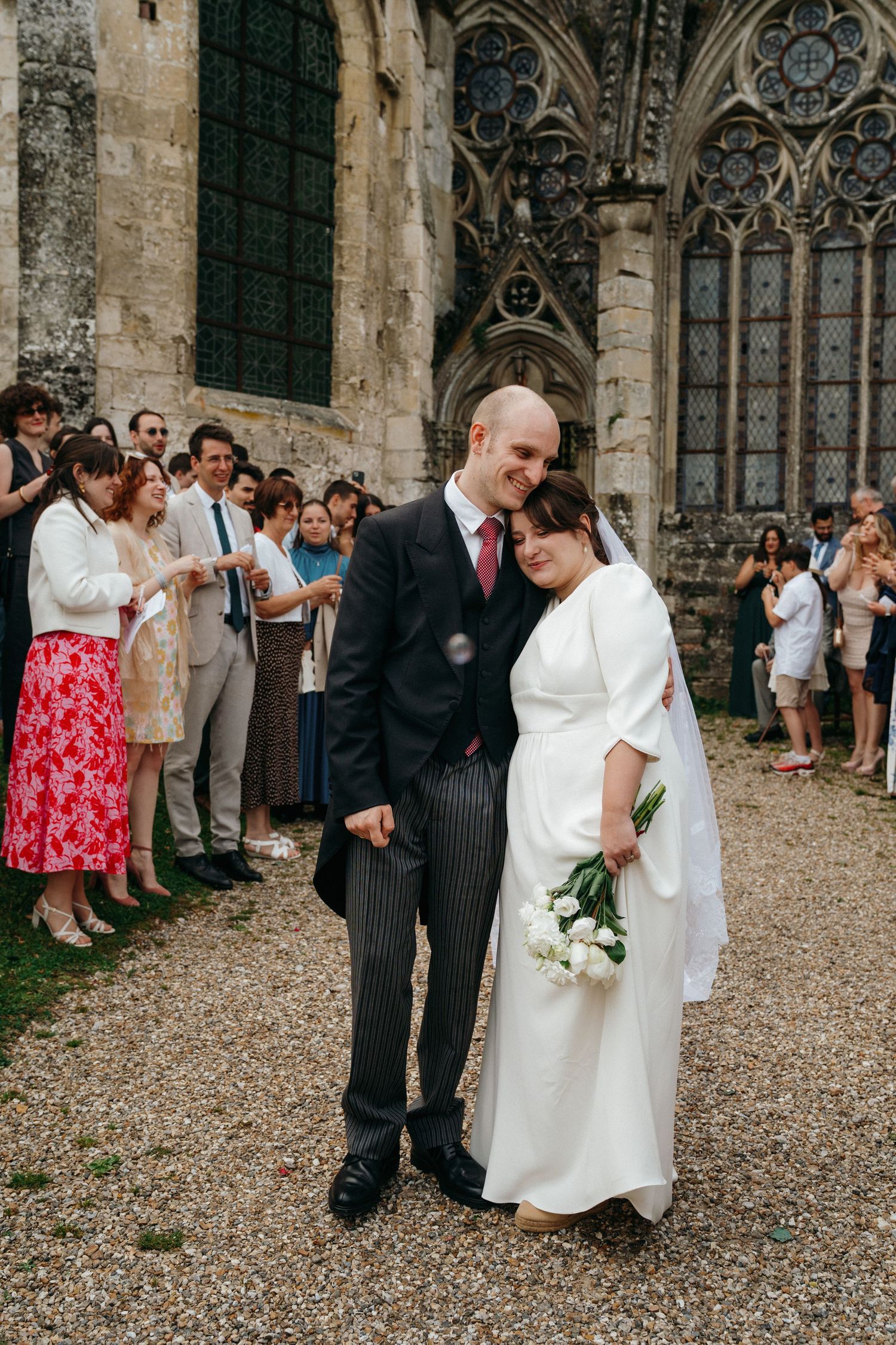 Newlywed couple smiling outside a Gothic cathedral surrounded by wedding guests throwing confetti.