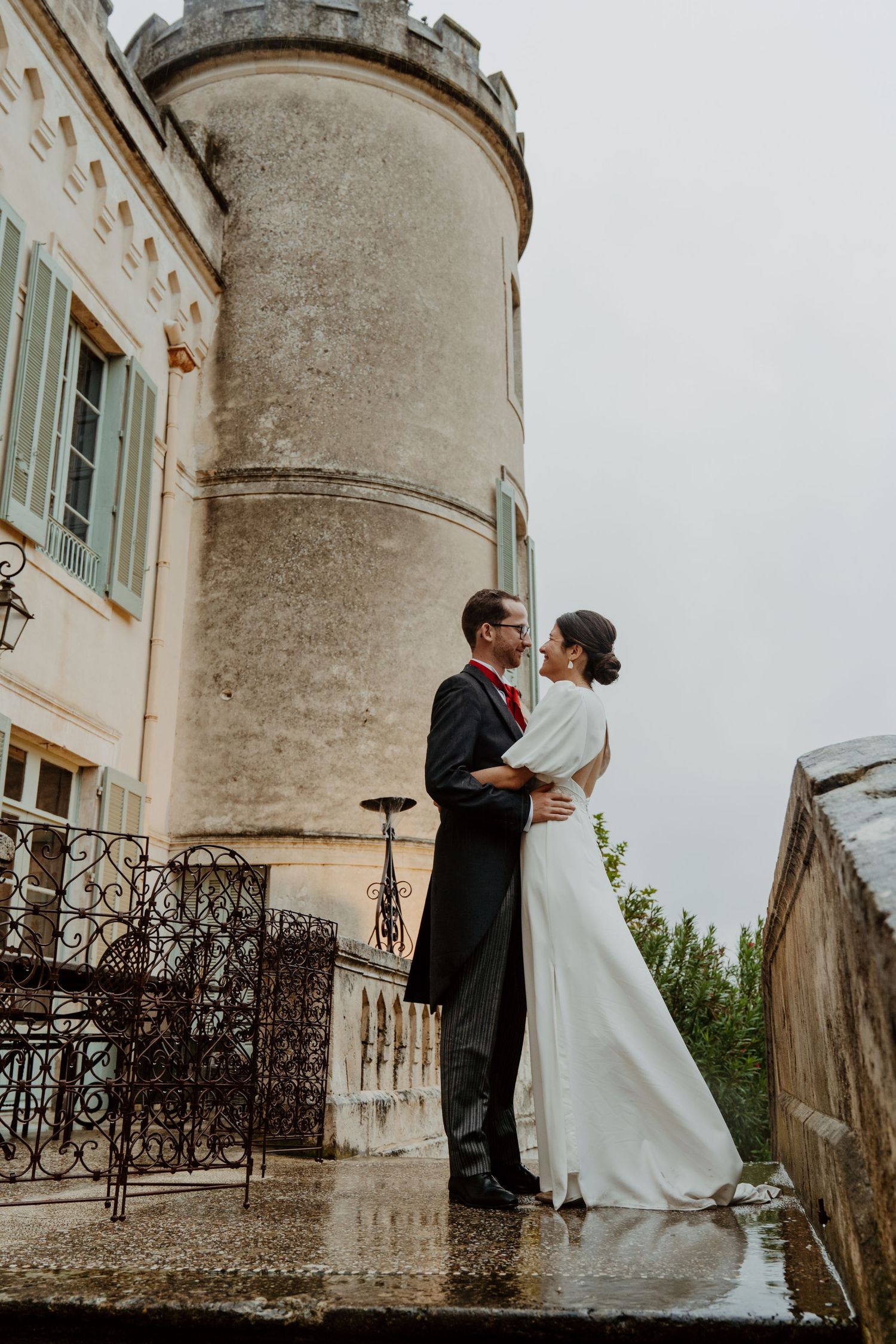 Elegant bride and groom embracing on wet terrace before historic French castle on cloudy day.