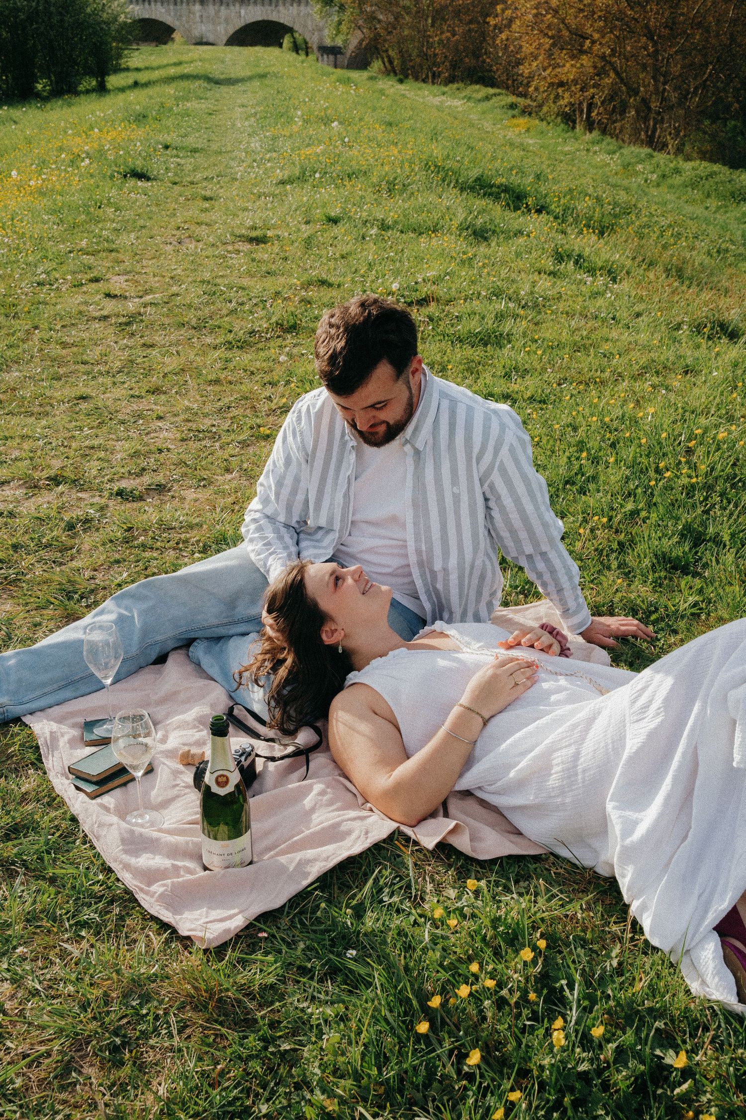 Couple relaxes on a blanket by a riverside meadow with champagne bottle and wine glasses nearby.