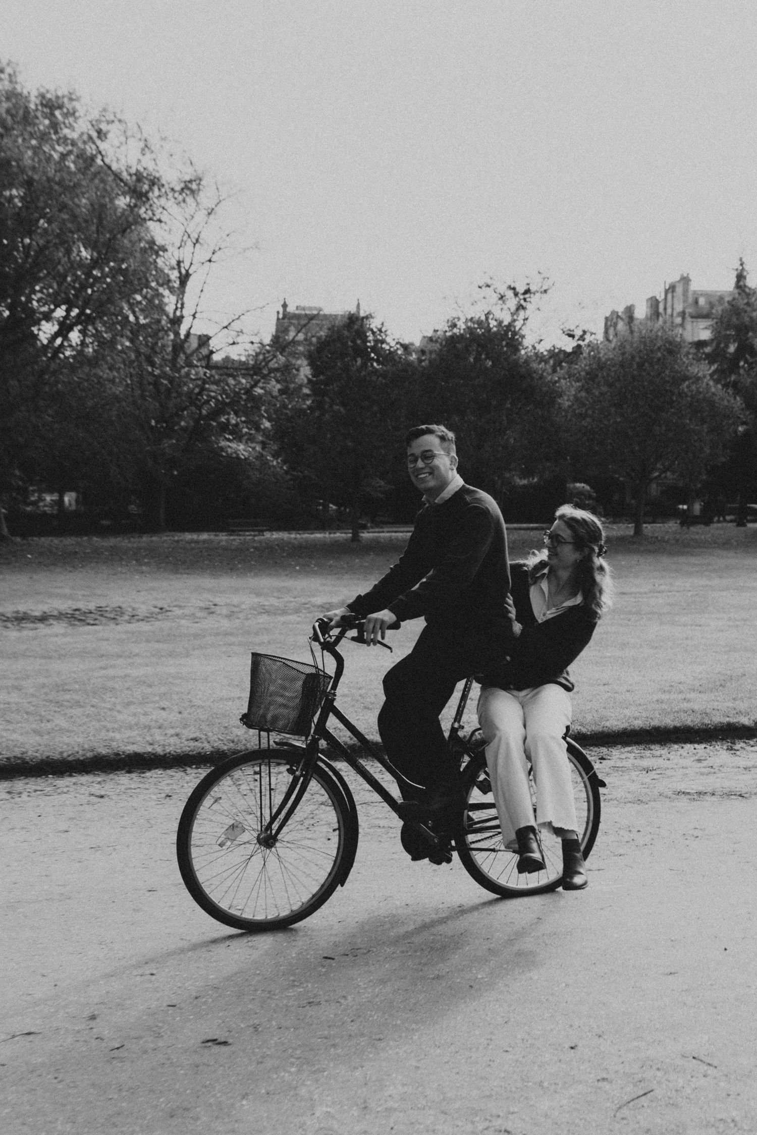 Black and white photo of a smiling couple riding a bicycle together through a park.