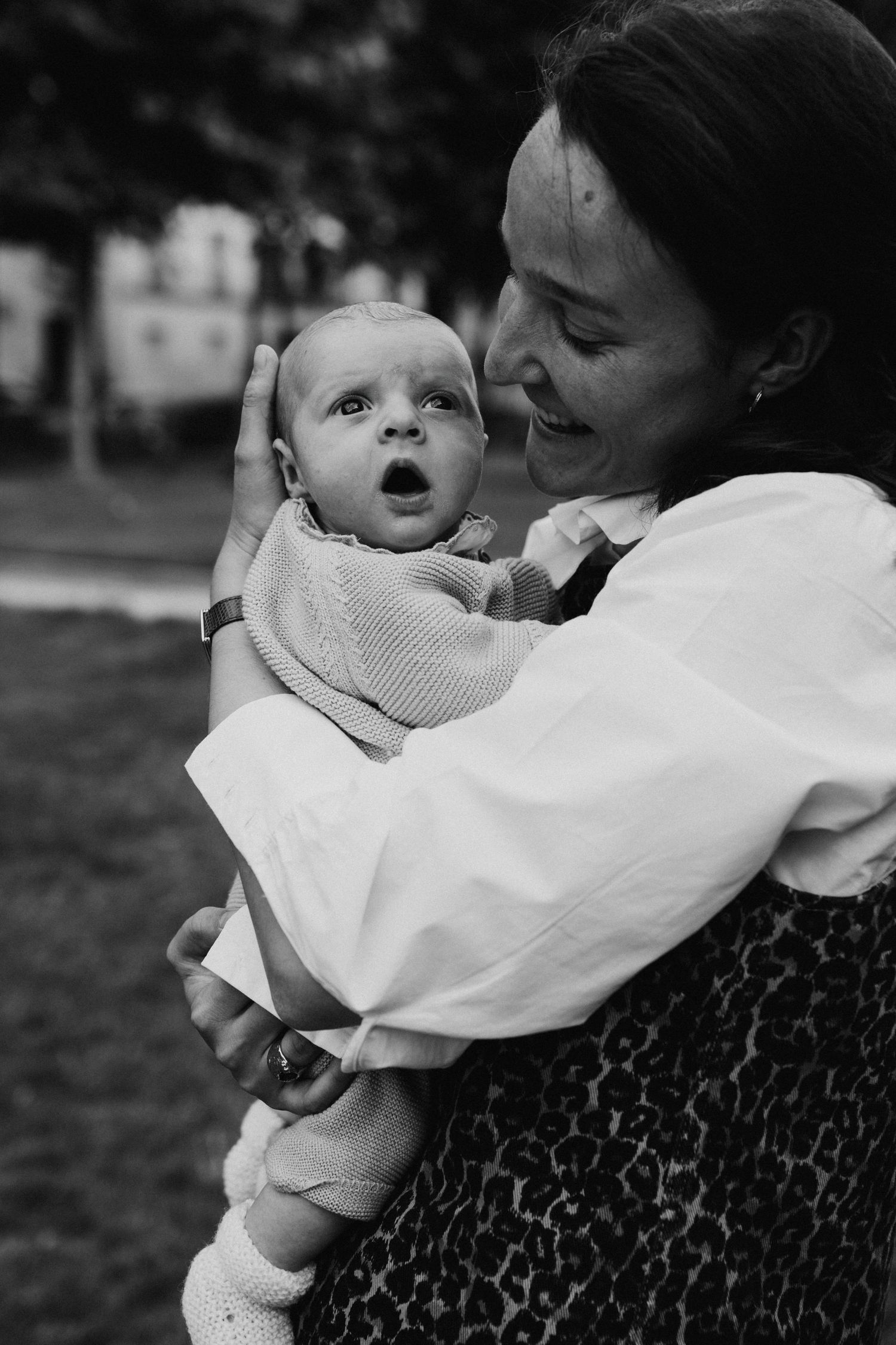 Black and white portrait of a smiling mother tenderly holding her wide-eyed newborn baby outdoors.