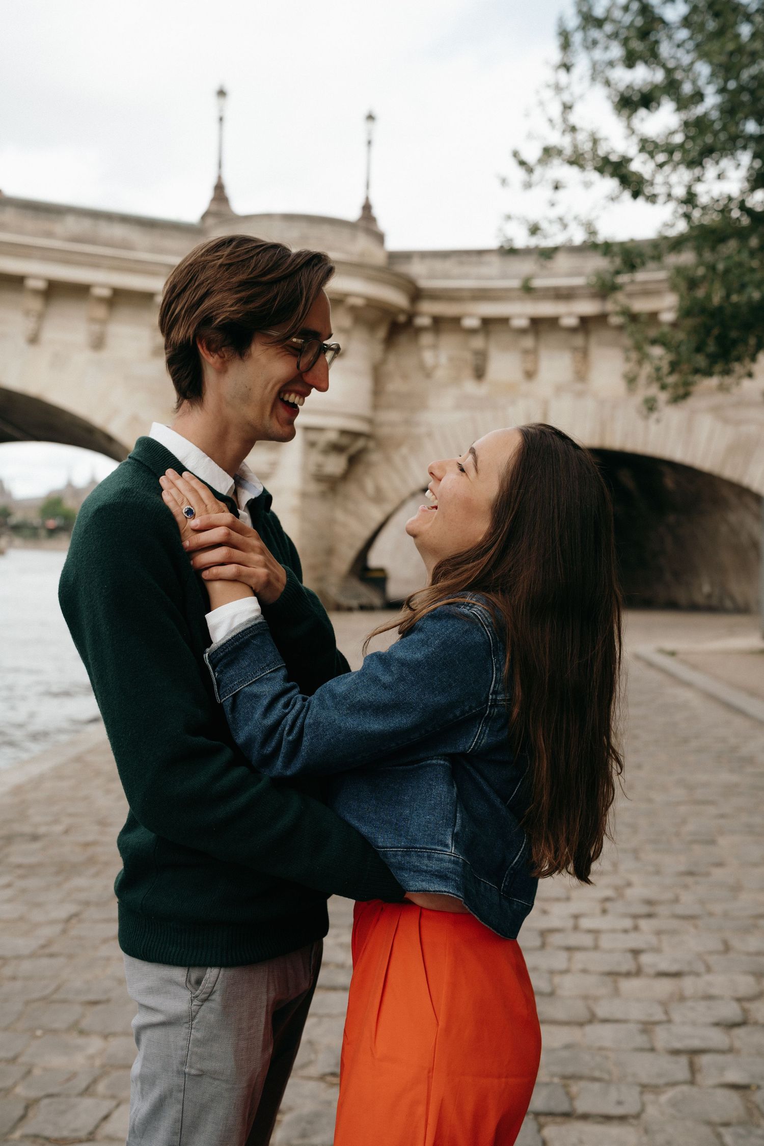 Happy couple embracing and laughing together on a cobblestone riverbank with a historic stone bridge in Paris.
