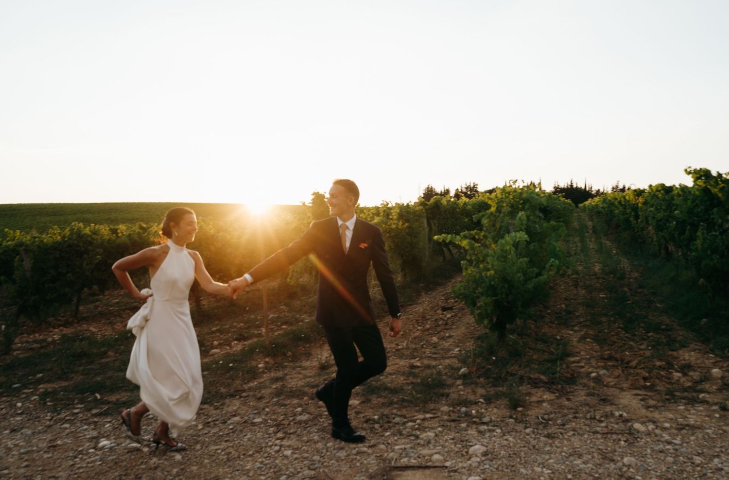 Bride and groom run hand-in-hand through sun-drenched vineyard rows at golden hour.