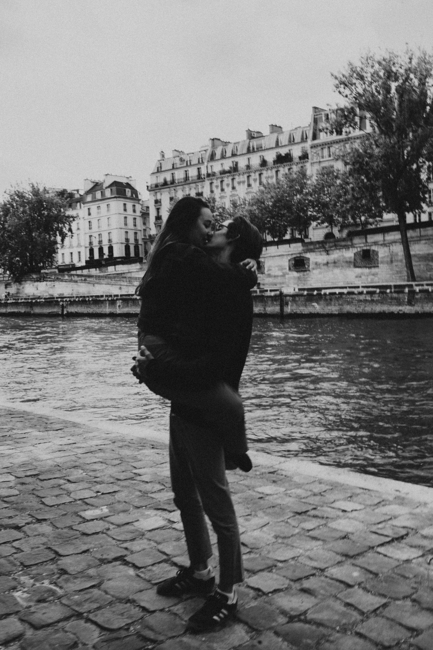 Romantic black and white photo of couple kissing by the Seine river with Parisian buildings in background.