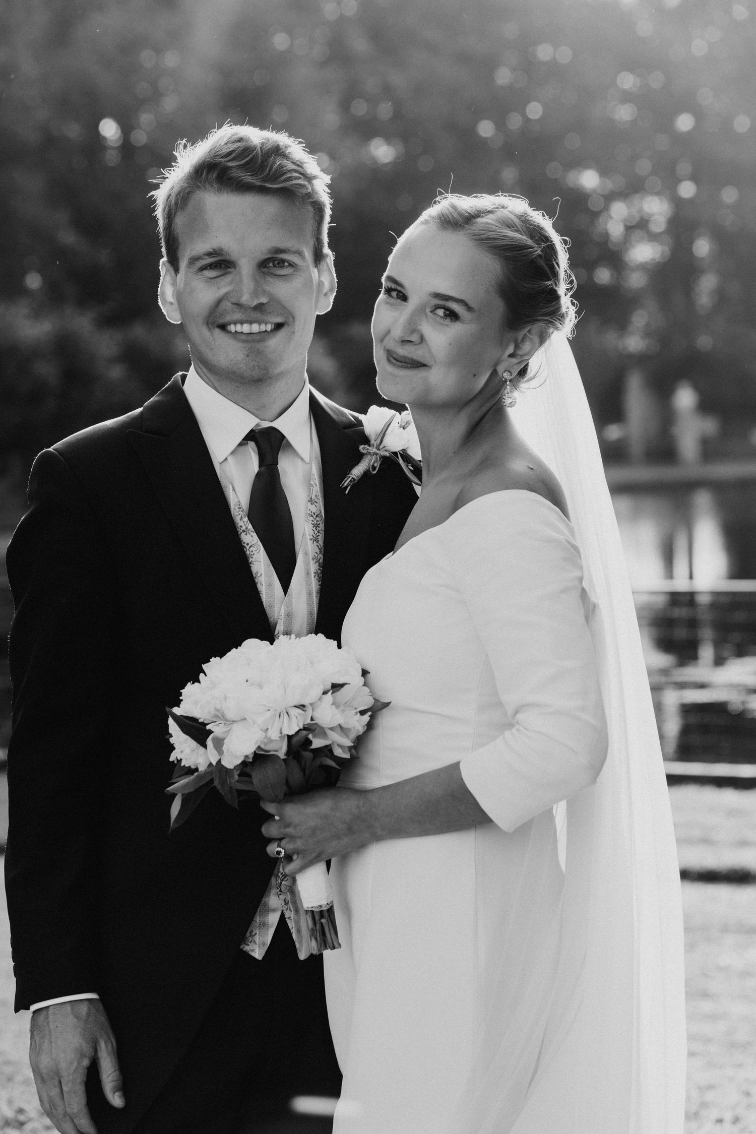 Black and white portrait of smiling newlyweds, bride holding peony bouquet wearing off-shoulder gown.