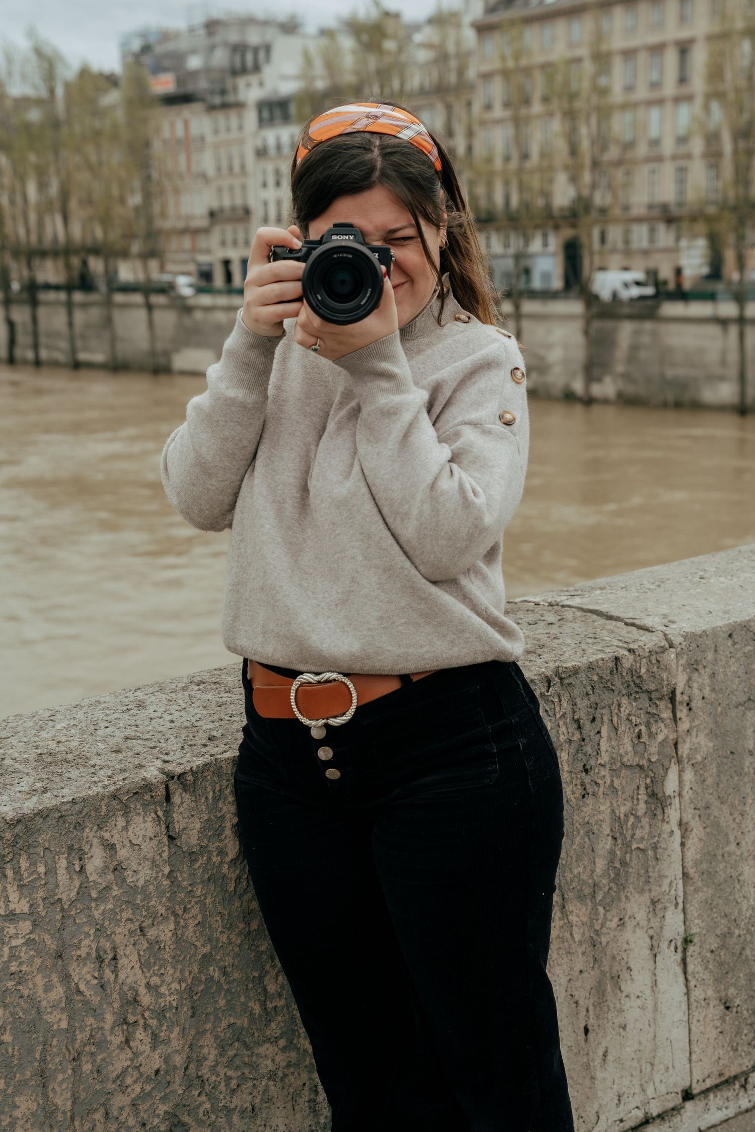 Female wedding photographer in casual attire shooting with camera on Parisian bridge.