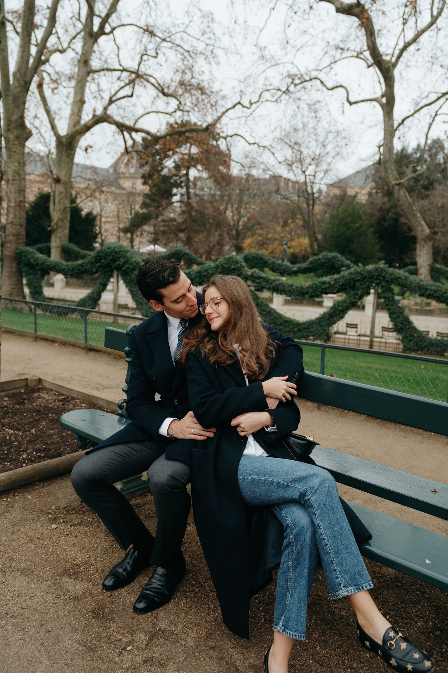 Un couple d'amoureux assis sur un banc dans un parc à Paris ; l'homme embrasse la femme sur la joue ; tous deux portent un manteau noirs.