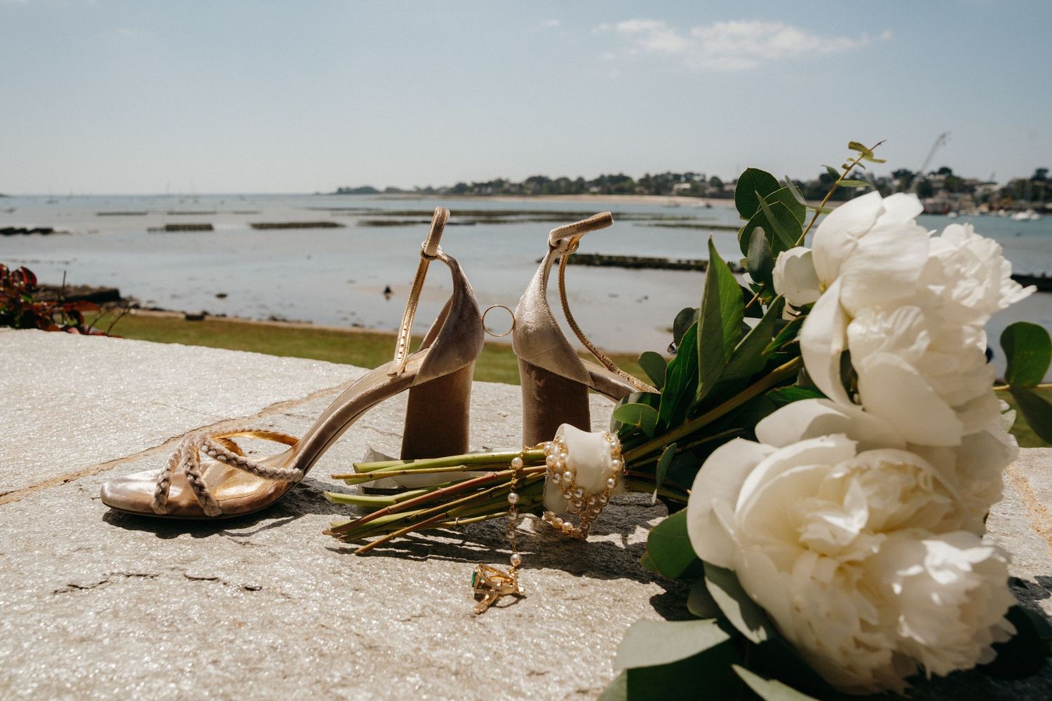 Elegant gold strappy wedding heels and white peony bouquet displayed on stone surface overlooking coastal water.