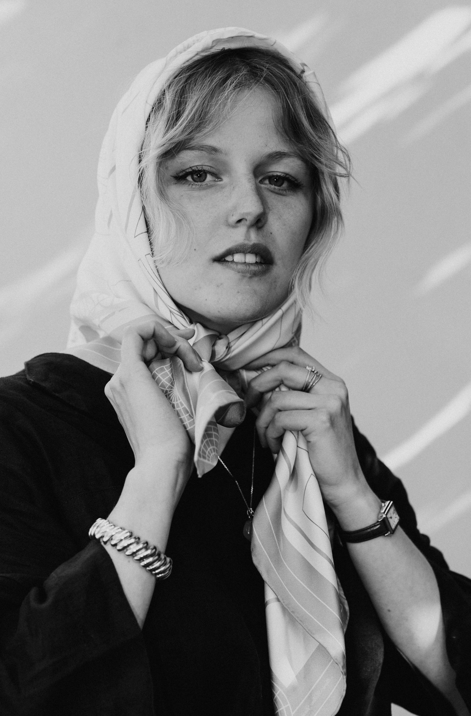 Black and white portrait of a young woman tying a white headscarf with freckled features.