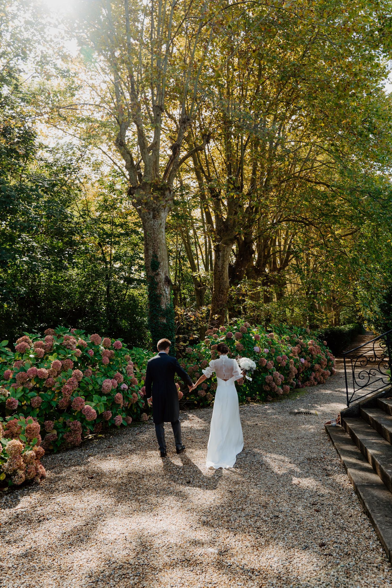 Bride and groom walk hand-in-hand along a gravel path lined with pink hydrangeas in a lush garden.