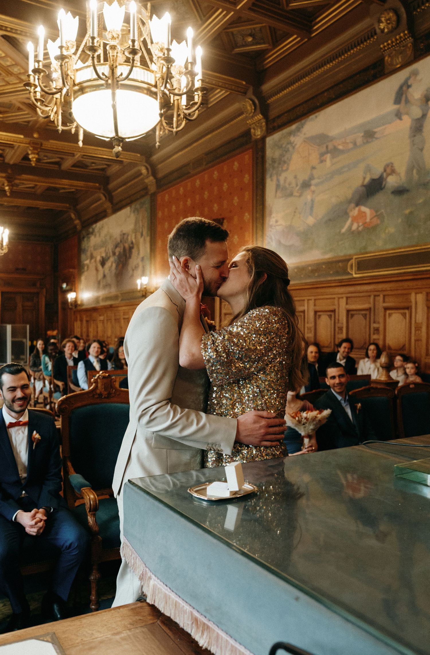 Couple shares a kiss at the altar in a grand wood-paneled hall with chandelier and guests seated behind them.