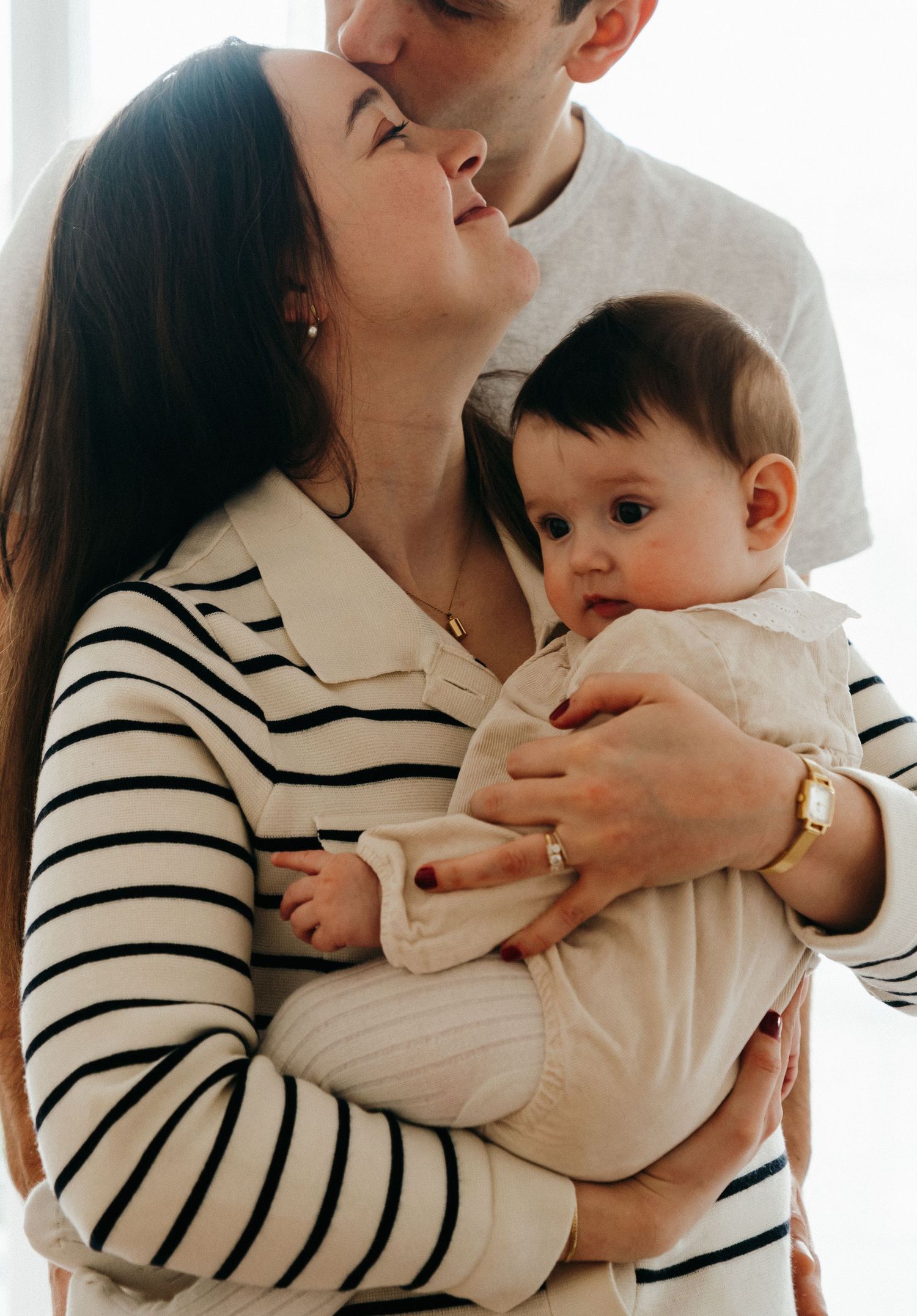 Young couple sharing a kiss while mother holds their dark-haired baby in a striped top.