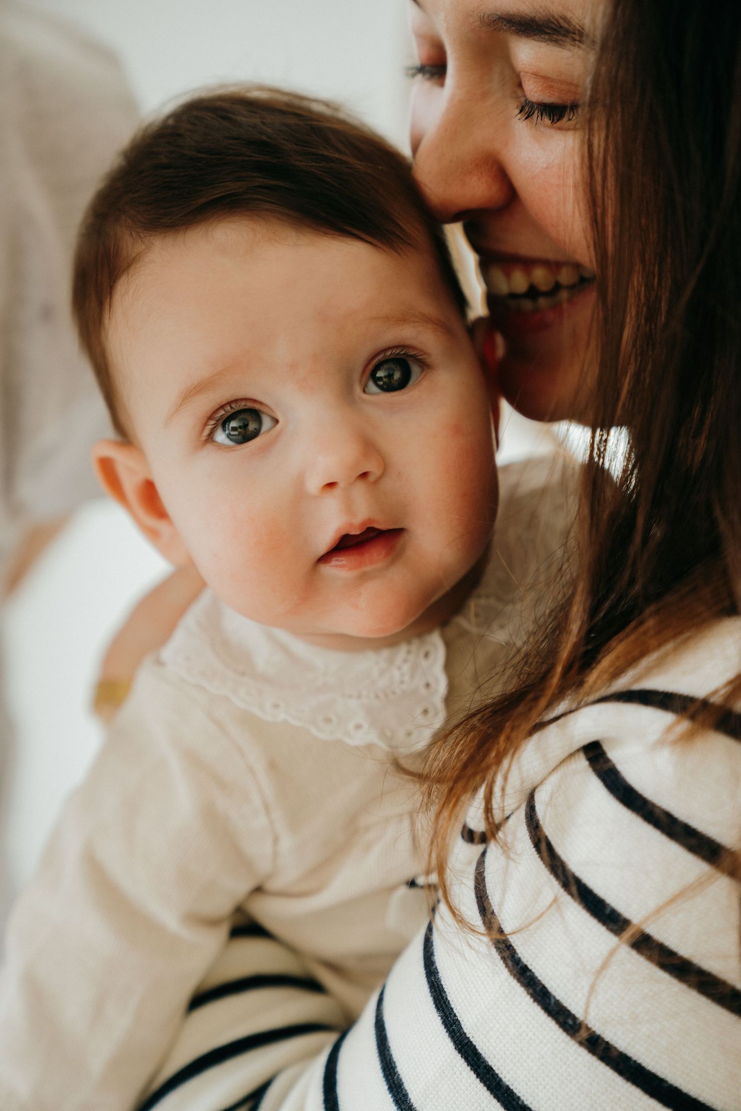 Close-up of a smiling mother kissing her adorable wide-eyed baby with dark hair.
