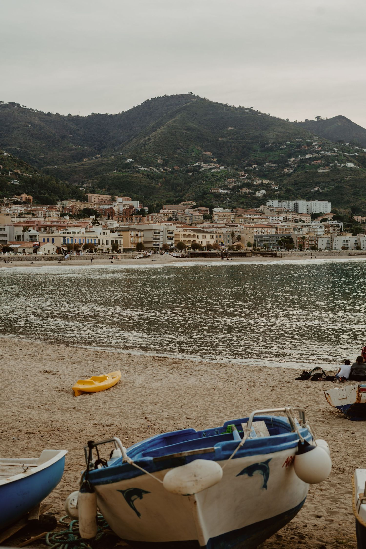 Scenic coastal town with boats on sandy beach, calm waters, and green mountains under overcast sky.