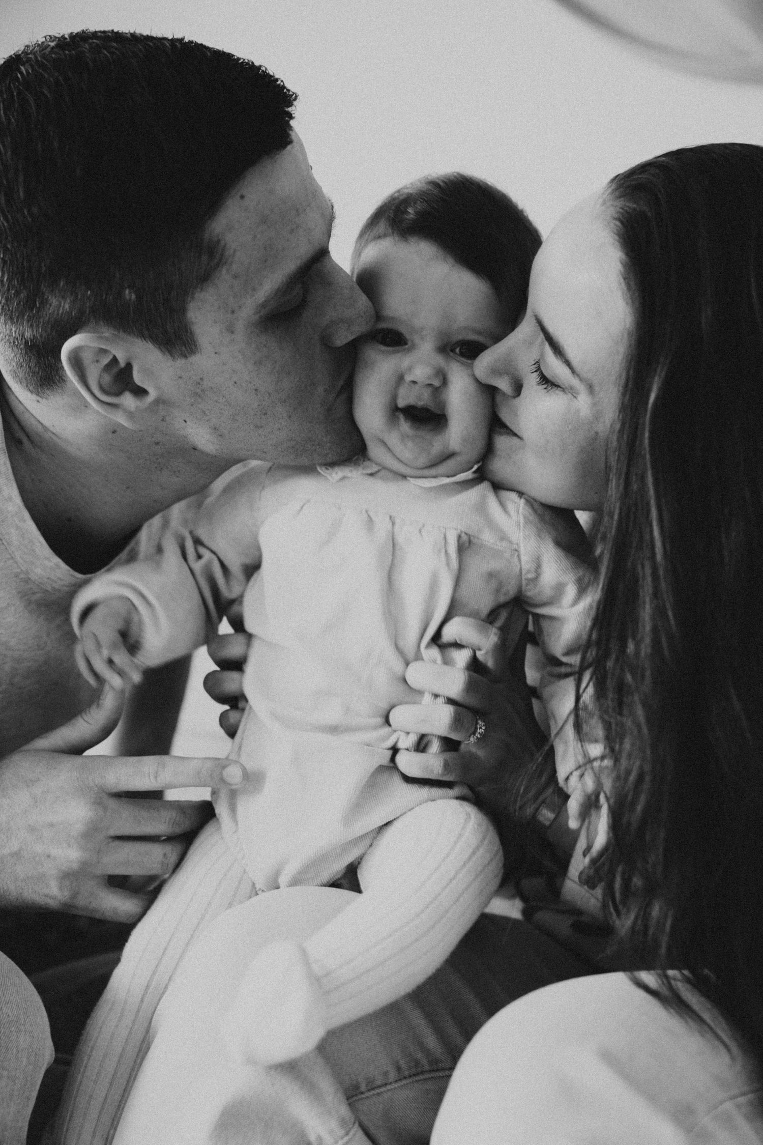 Photo en noir et blanc montrant des parents embrassant simultanément leur bébé, qui rit, sur les deux joues.