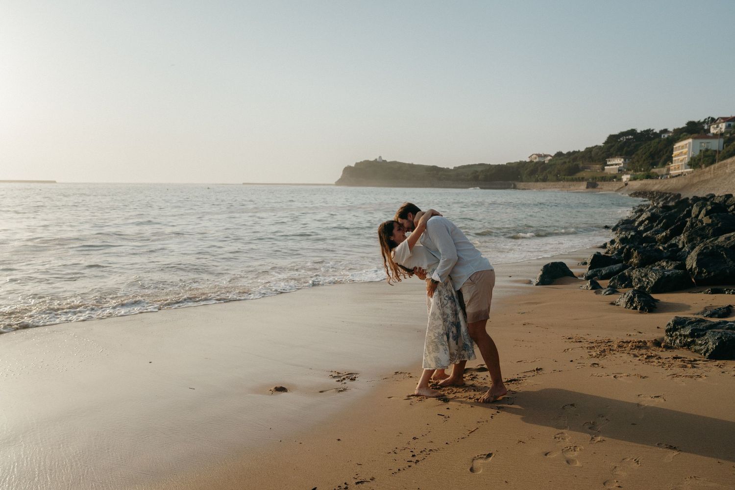 Couple shares a romantic kiss on a sandy beach at sunset with coastal cliffs in background.