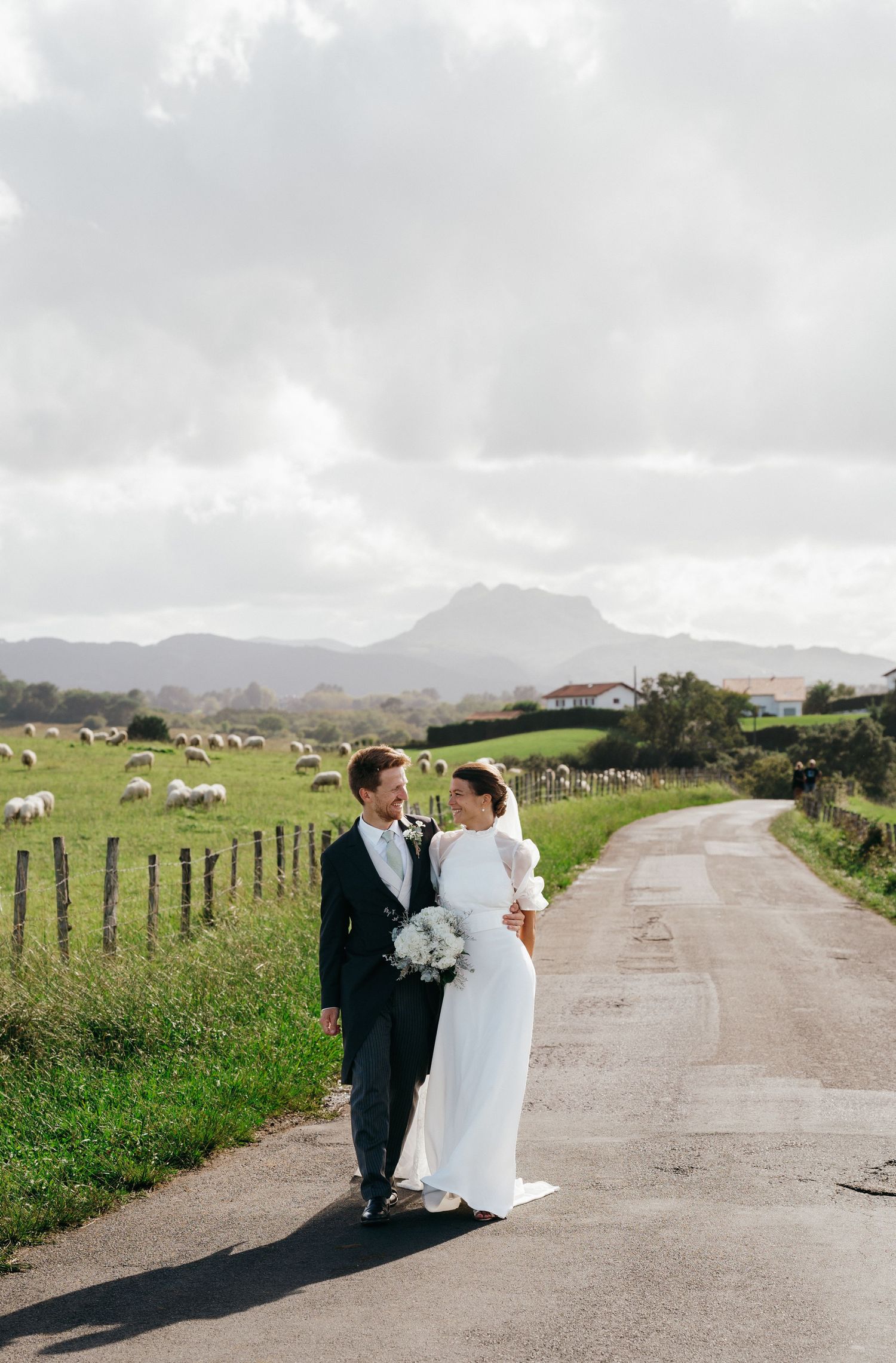 Bride and groom sharing a romantic moment on a rural road with sheep grazing and mountains in the background.