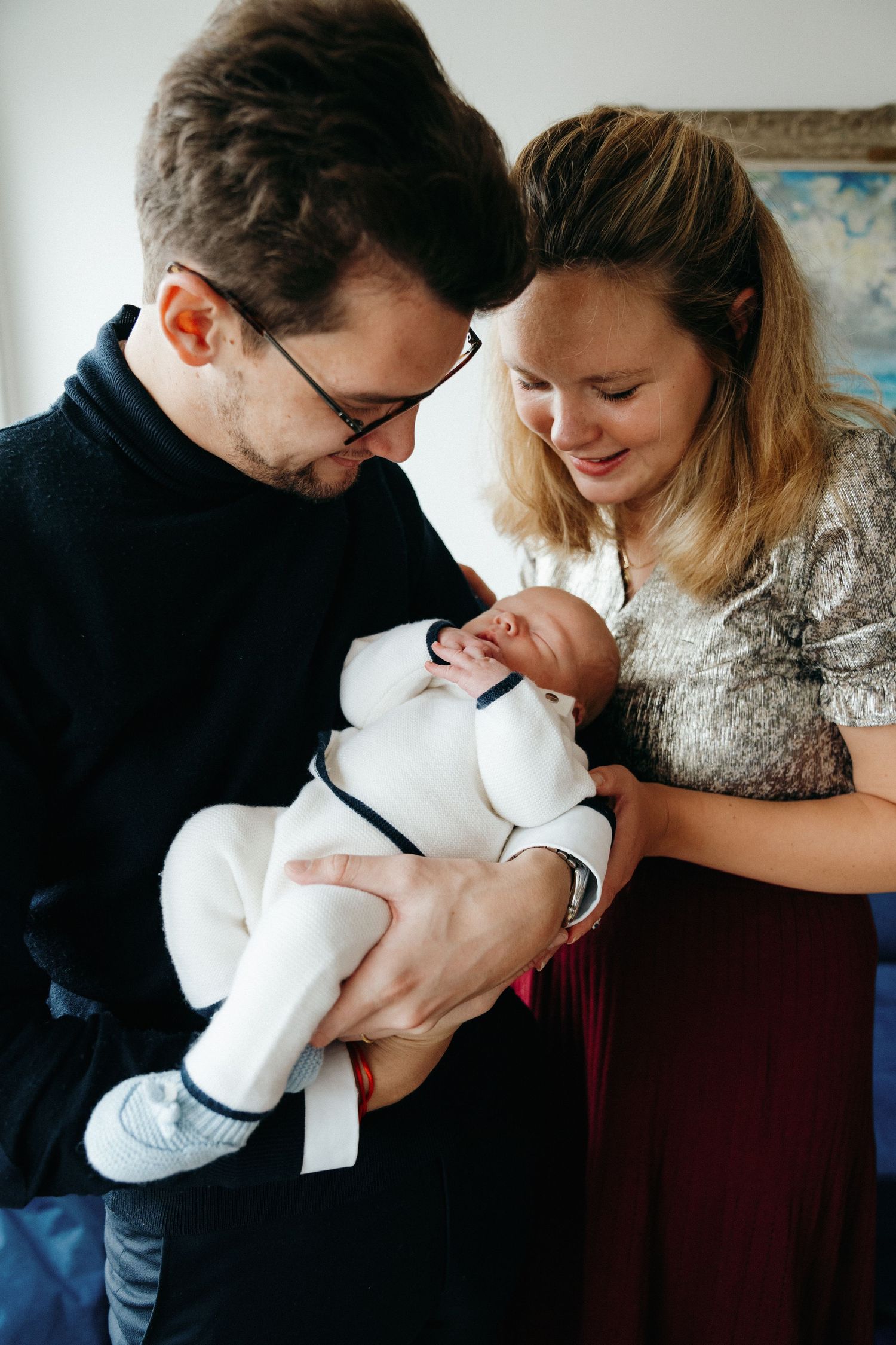 New parents in cozy home lovingly gazing at their newborn baby dressed in white outfit.