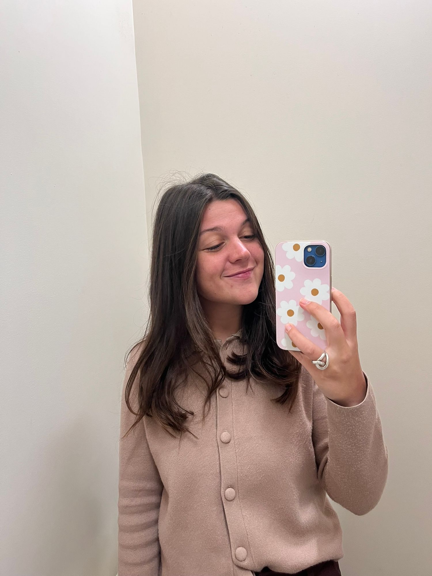 Young brunette woman taking a mirror selfie in a beige cardigan, holding a pink polka dot phone case.