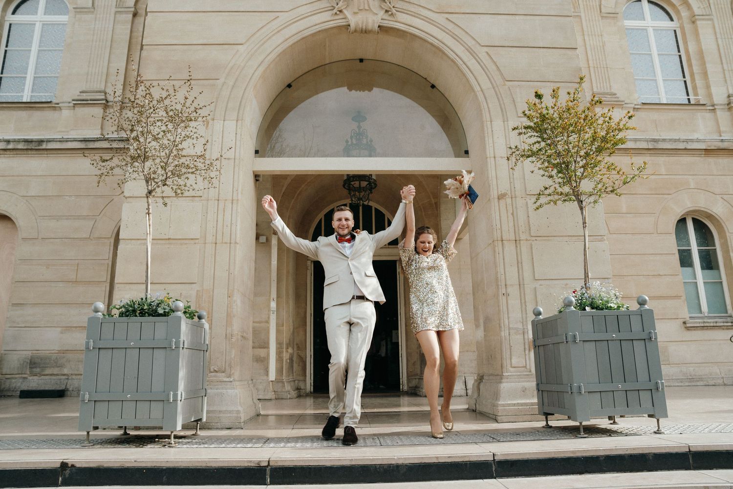 Joyful newlyweds jumping with raised hands outside a grand stone building after their wedding ceremony.