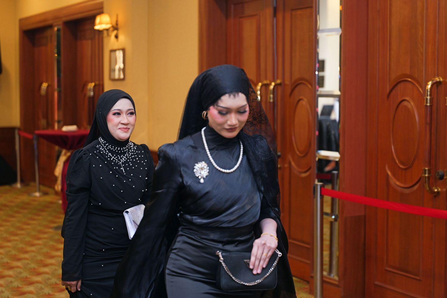 Two Malaysian women in elegant black attire walking through a formal event venue with red rope barrier.