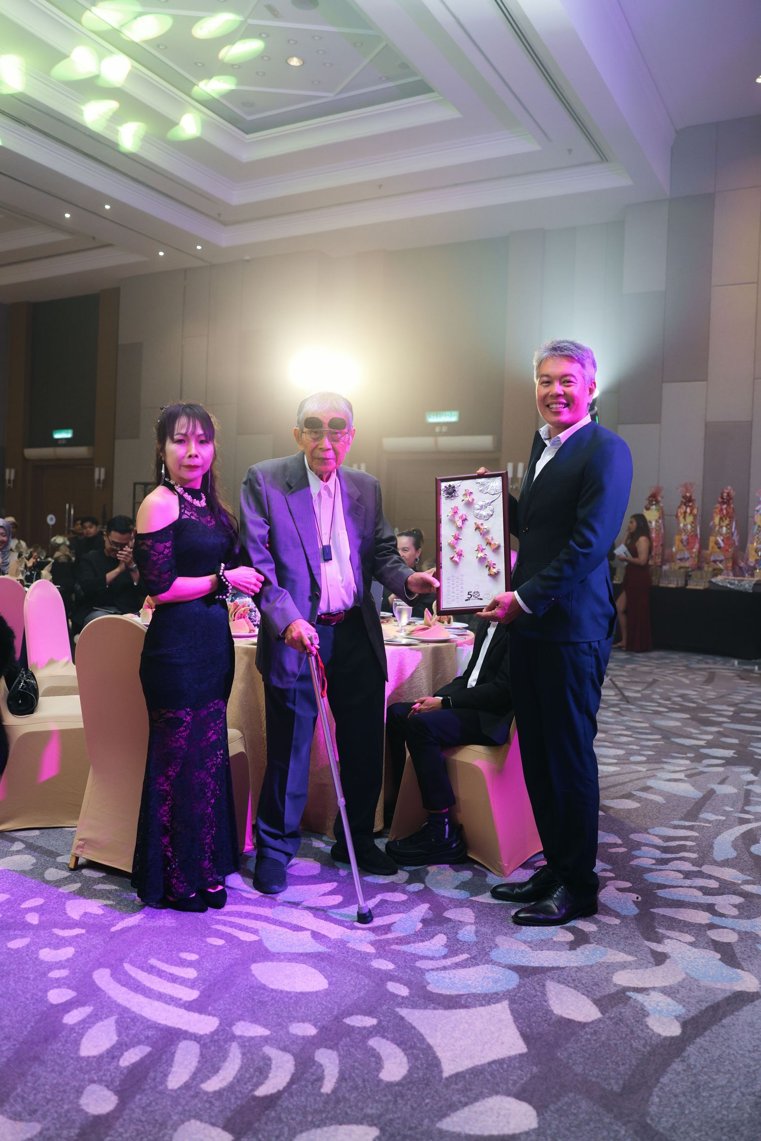 Three elegantly dressed guests pose together at a formal banquet hall event, holding a framed artwork.