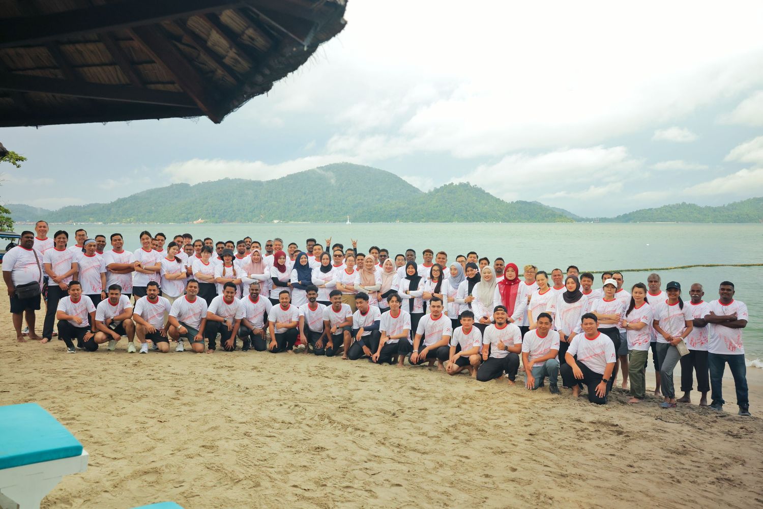 Large group of people in white shirts posing together on a sandy beach with mountains and calm water in the background.