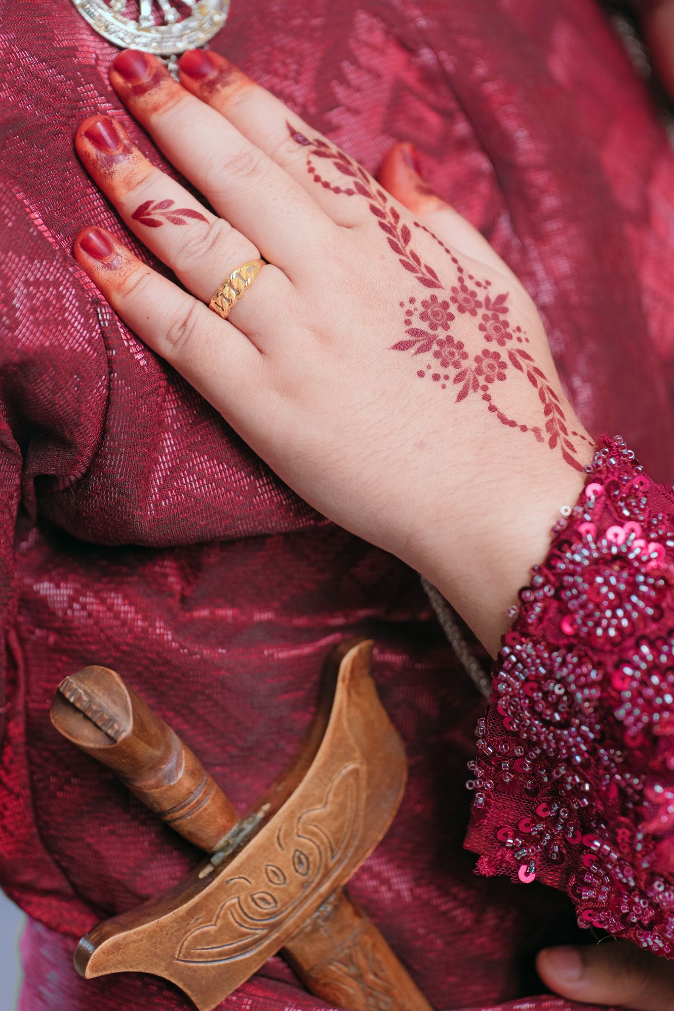 Hand adorned with henna design and gold ring rests on burgundy fabric near wooden decorative handle.