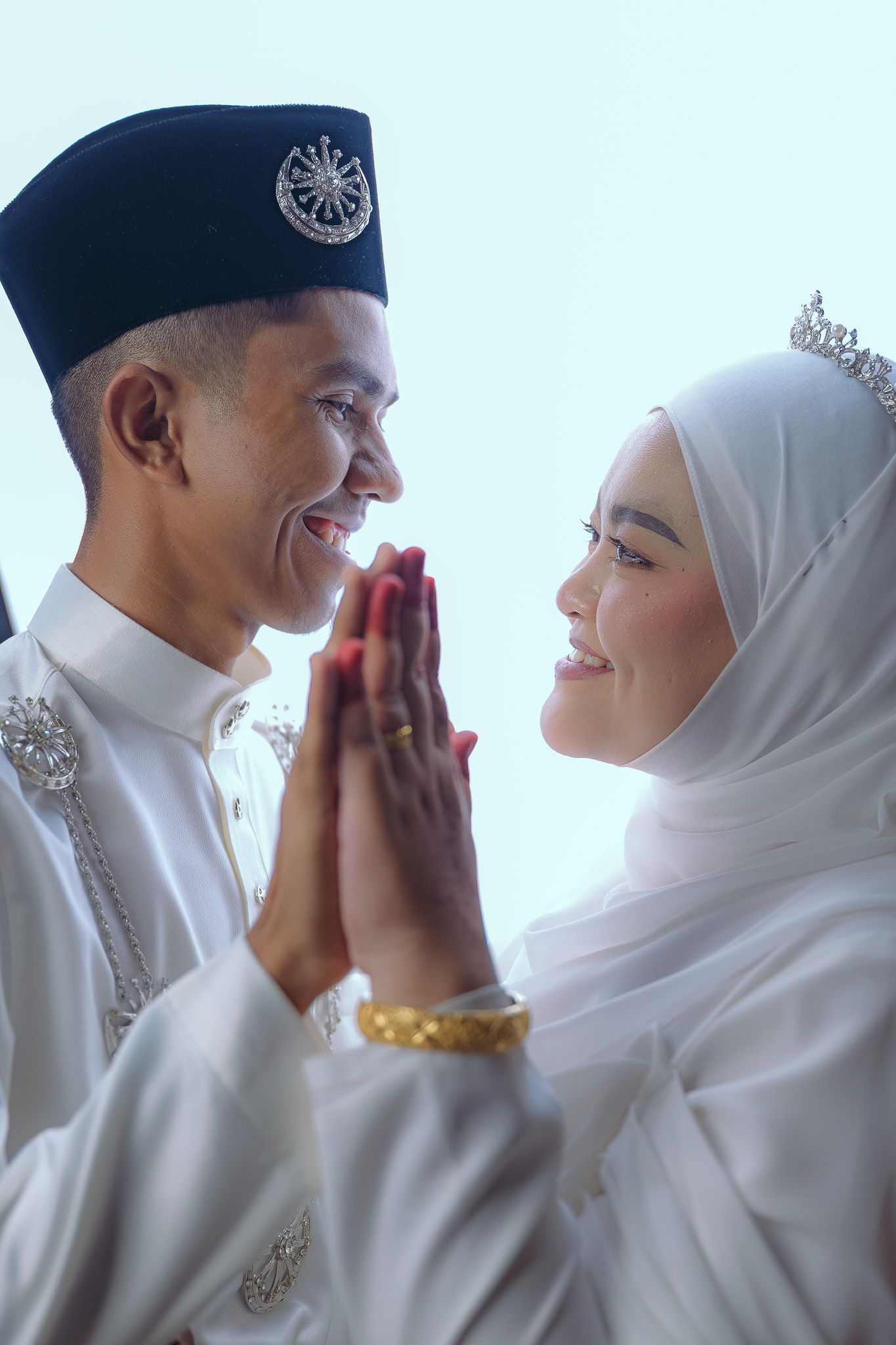 Muslim couple in white traditional wedding attire pressing palms together in joyful moment.