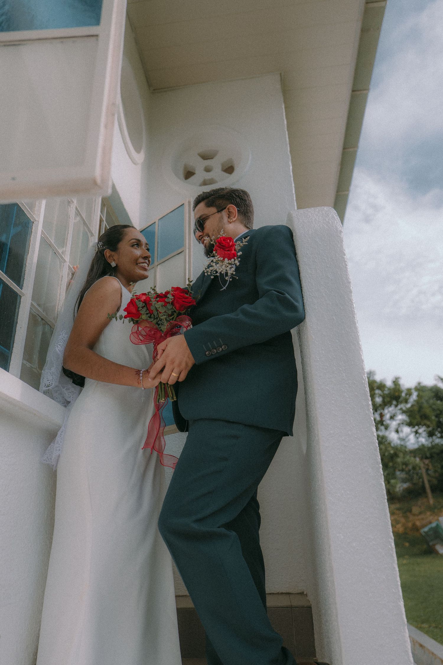 A couple shares a joyful moment with red flowers on a white porch with classical architecture.