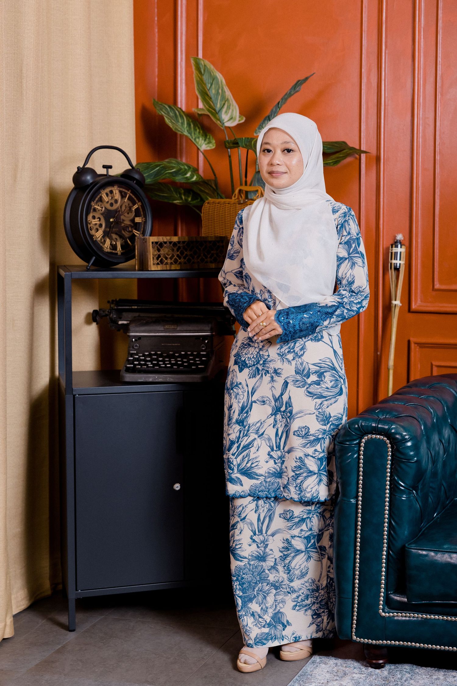 Person wearing white hijab and blue floral dress sits on navy velvet chair in room with wooden cabinets.