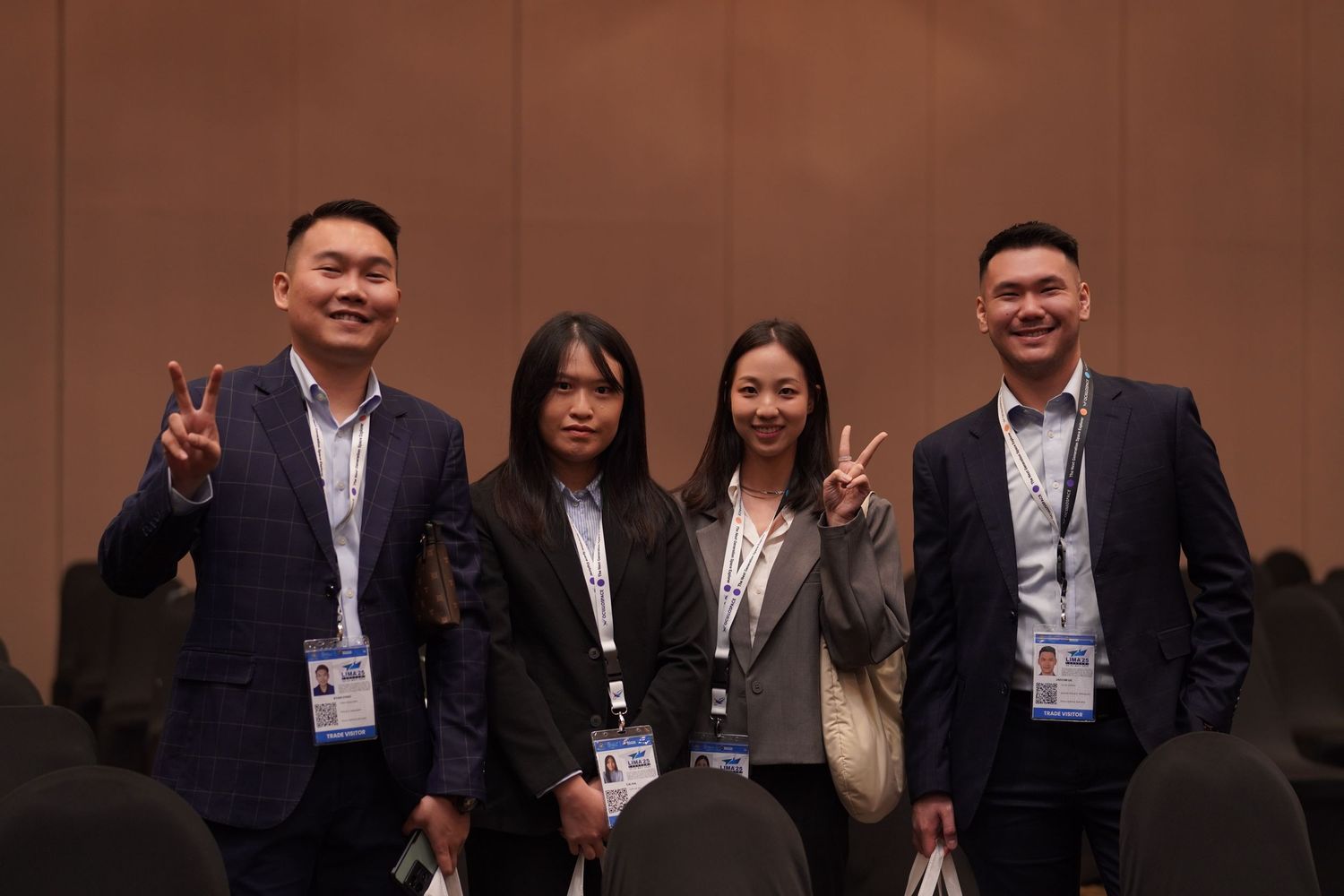 Four people in business attire pose together making peace signs at a professional conference.