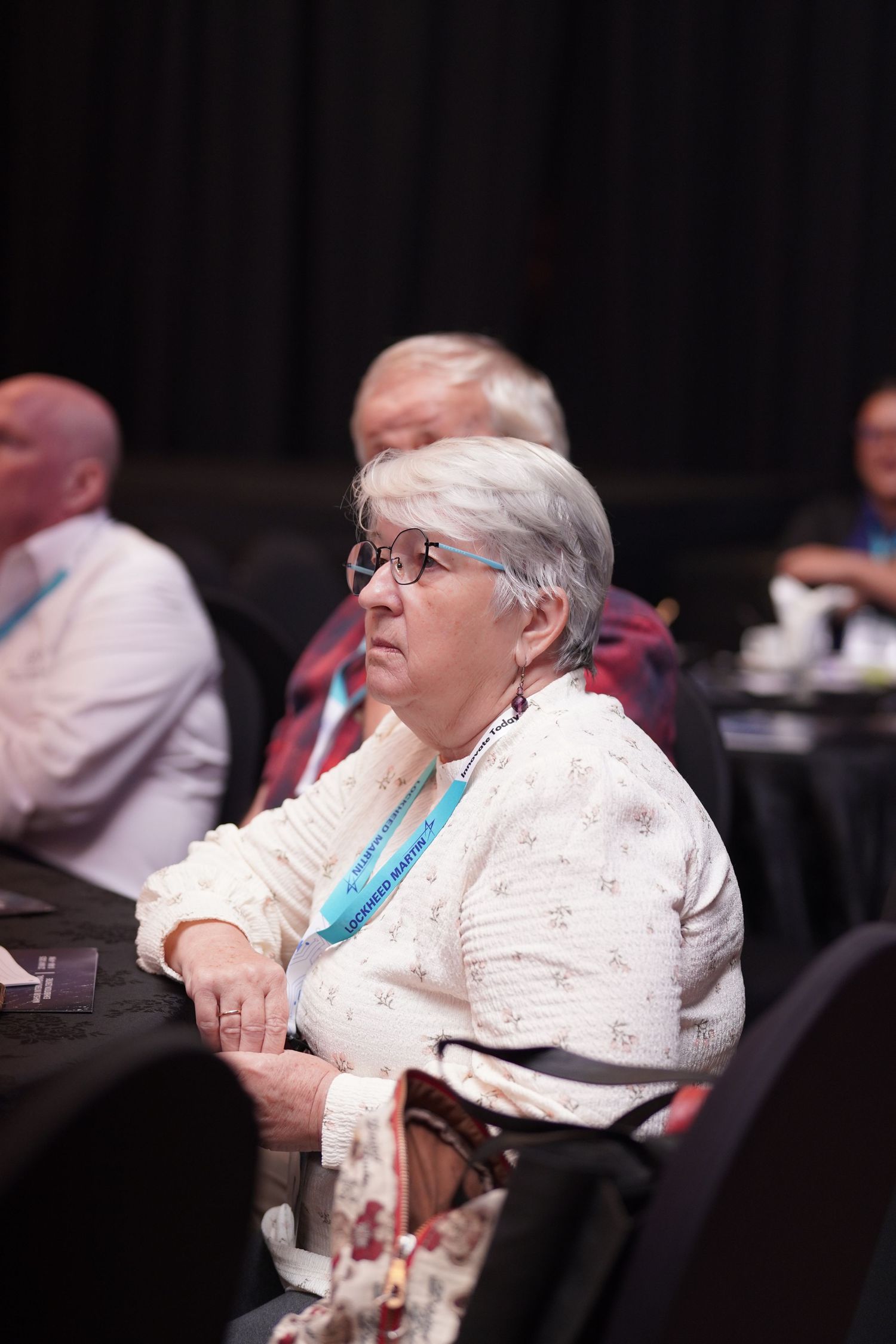 Conference attendee in white sweater and glasses sits attentively during a seminar session.