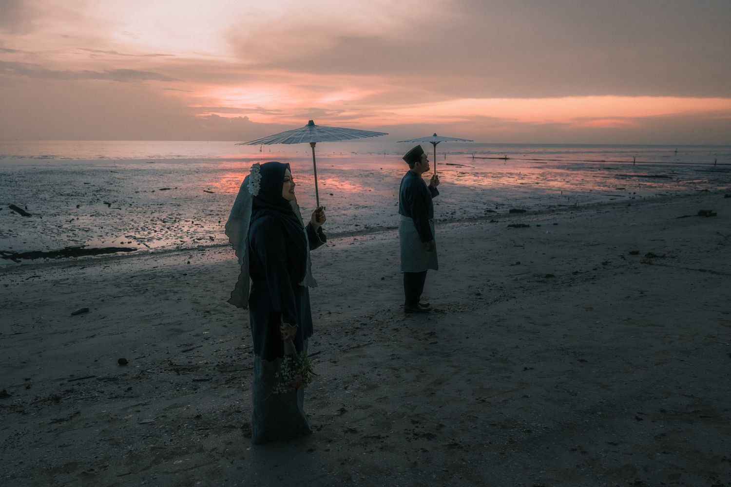 Silhouetted figures stand on a beach at sunset holding fishing rods against a dramatic orange sky.