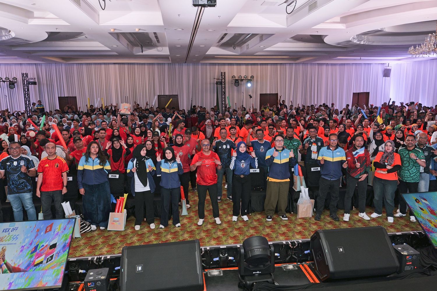 Large crowd of participants in colorful shirts gathered in a grand ballroom for a corporate or sports event.